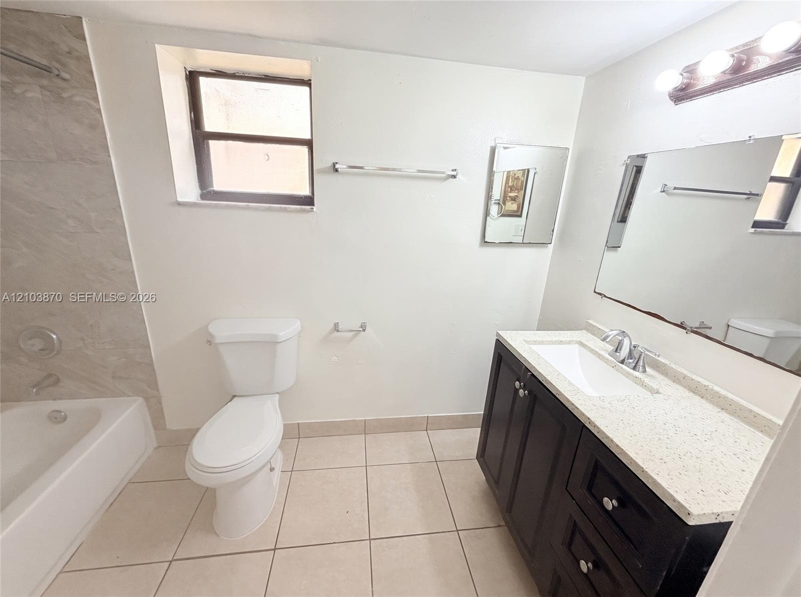 Bathroom featuring a dark wood-finish vanity with a light-toned solid surface countertop, an integrated rectangular sink, large format tile flooring, and a bathtub with neutral-tone wall tile surround