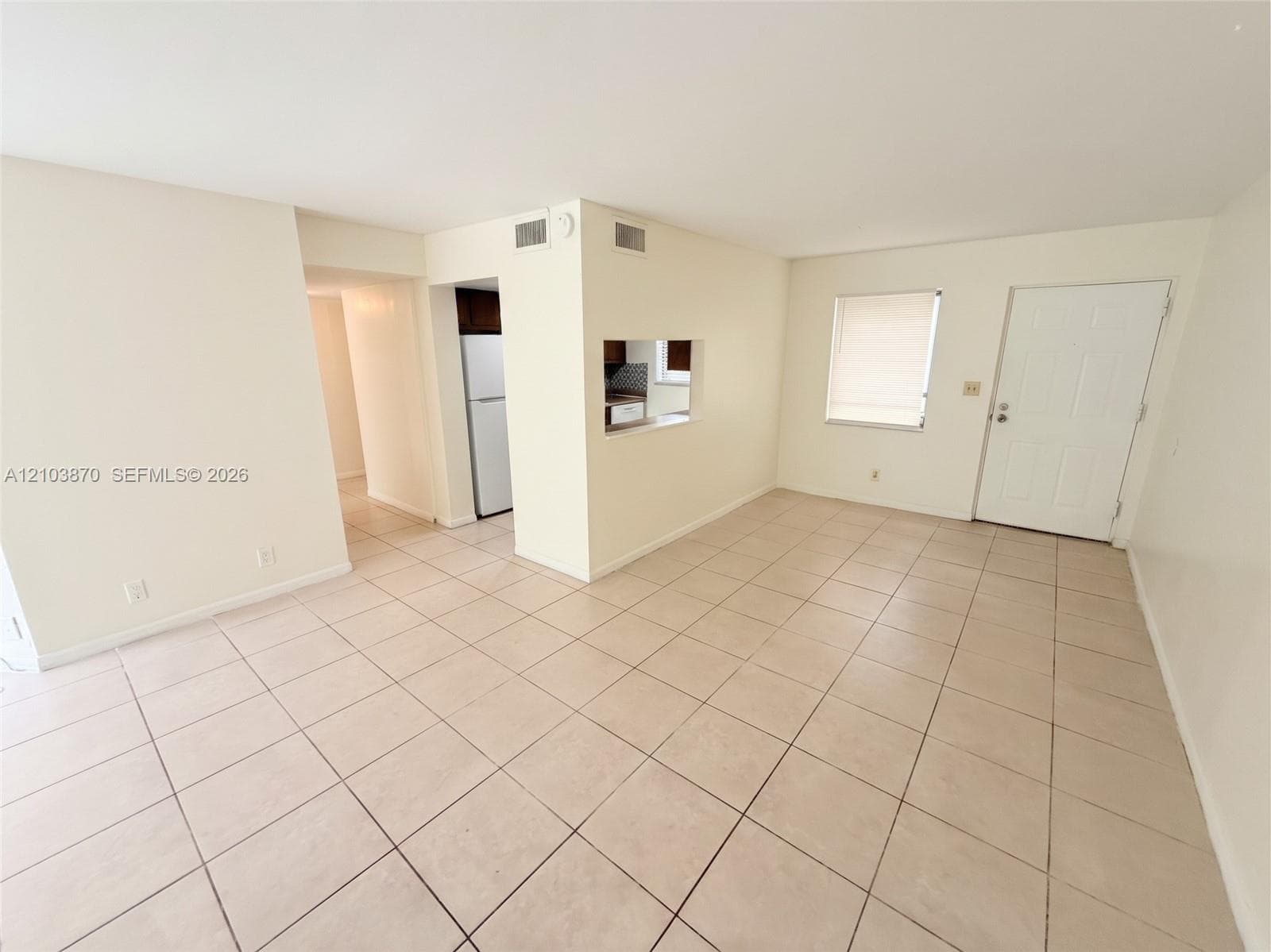 Open living space featuring neutral ceramic tile flooring, a window with horizontal blinds, and a pass-through opening to the kitchen area