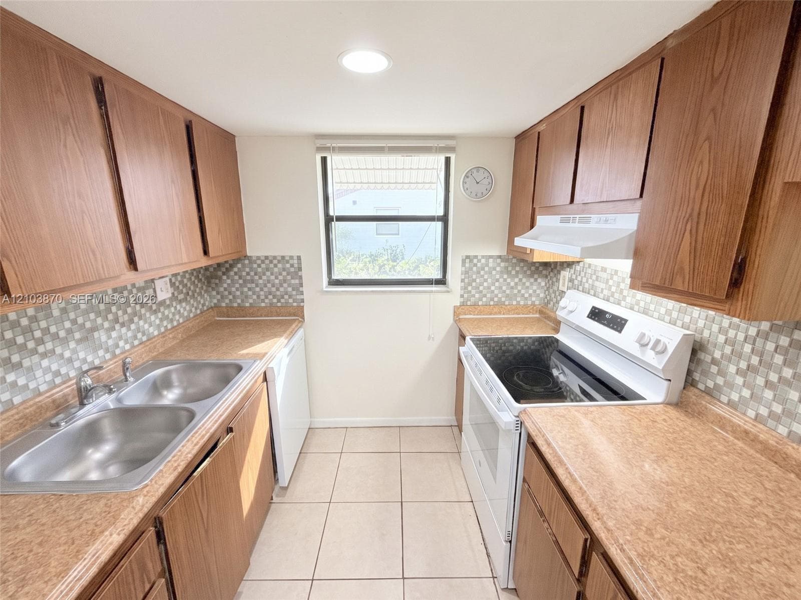 Kitchen featuring wood-finish cabinetry, neutral-toned laminate countertops, a mosaic tile backsplash, tile flooring, and a stainless steel double-basin sink