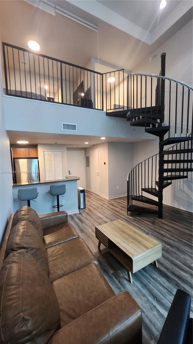 High-ceiling living area featuring wood-finish flooring, a metal spiral staircase with matching railing, and a kitchen breakfast bar with integrated stainless steel appliances