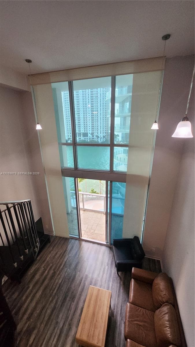 Double-height floor-to-ceiling windows, wood-finish flooring, metal staircase railing, and multiple pendant light fixtures