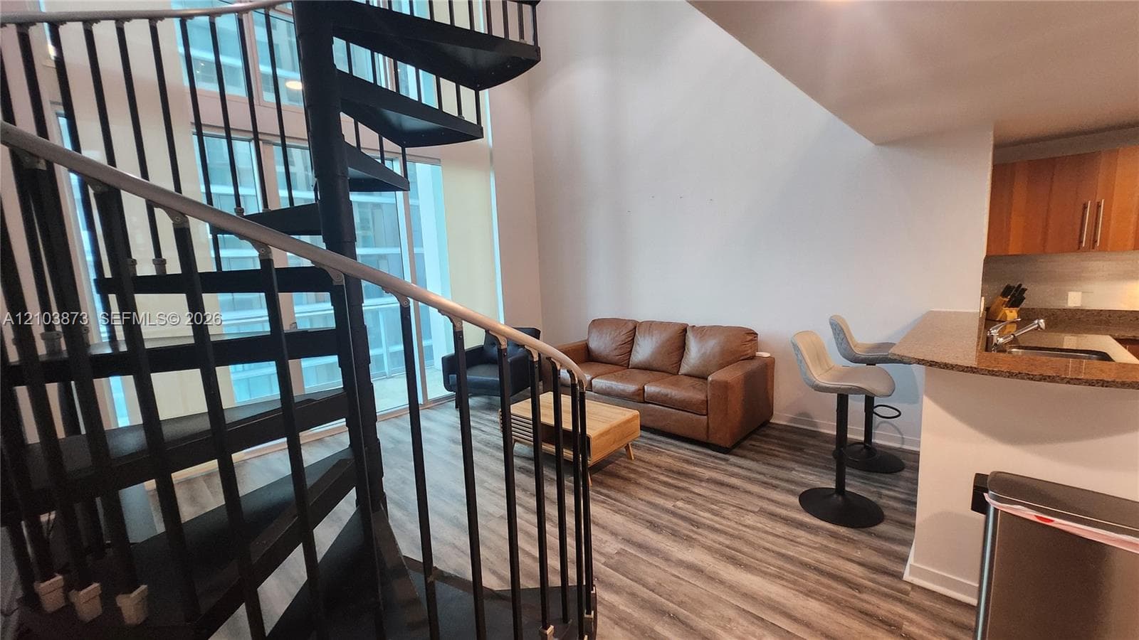 Black metal spiral staircase with wood treads, soaring cathedral ceilings, expansive floor-to-ceiling windows, wood-finish flooring, and a kitchen breakfast bar with stone-surfaced countertop