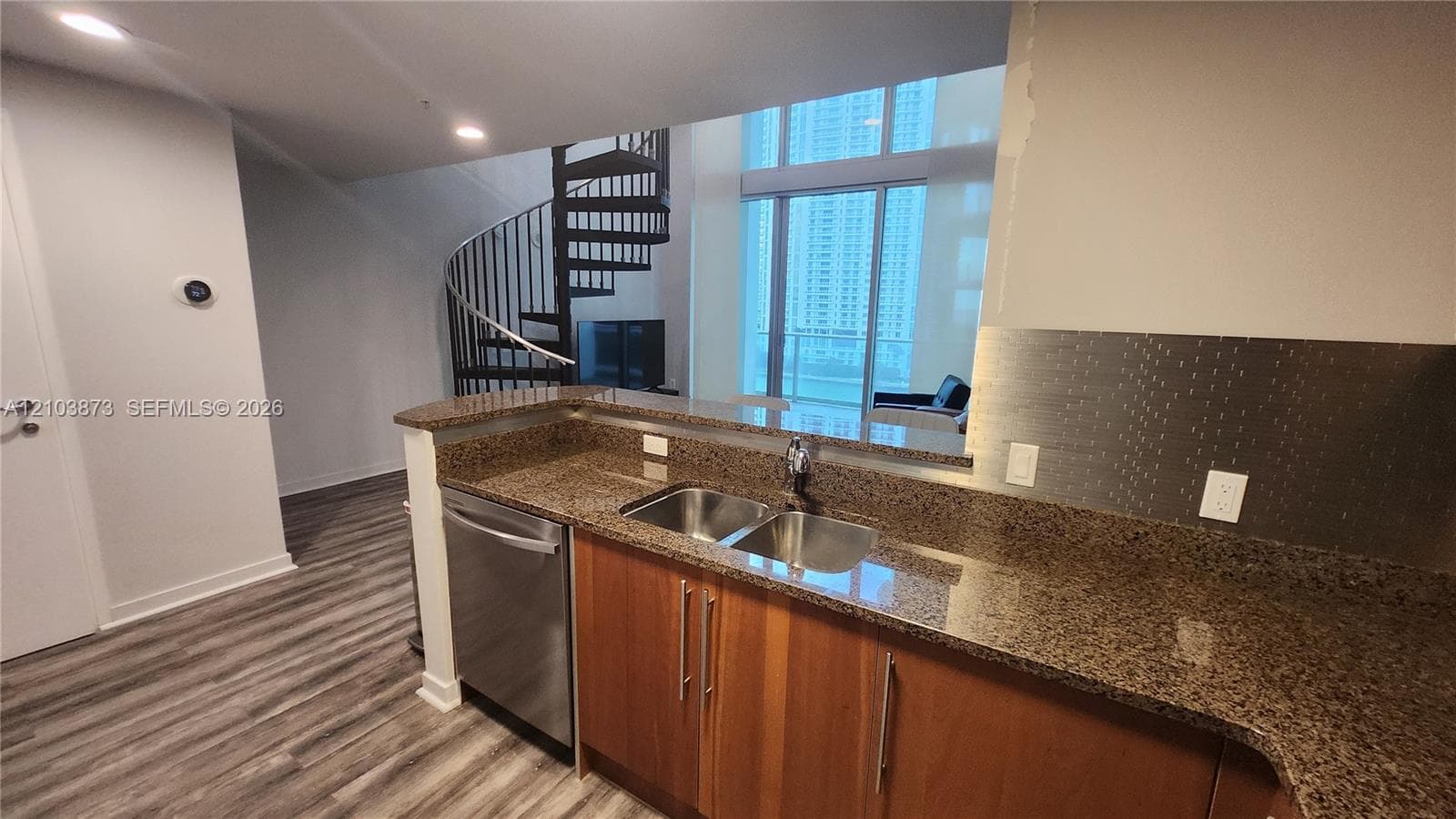 Kitchen featuring stone countertops, stainless steel dual-basin sink, wood-finish cabinetry, and stainless steel dishwasher, complemented by a metal spiral staircase and floor-to-ceiling windows