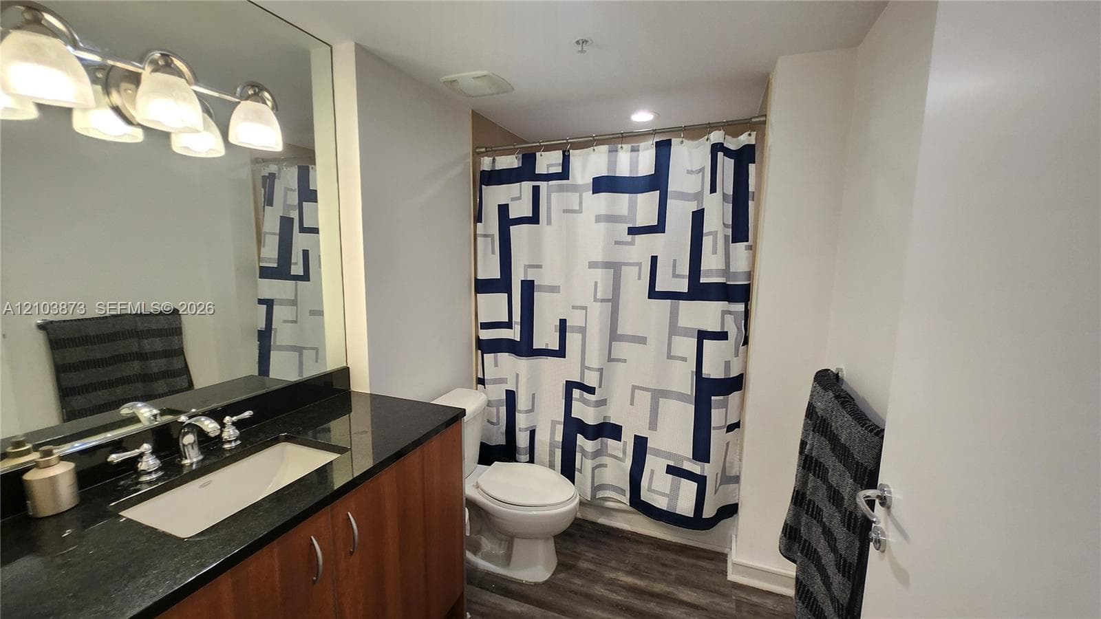 Bathroom featuring a dark stone countertop, integrated sink, wood-finish vanity cabinetry, four-light vanity fixture, and full-width wall mirror