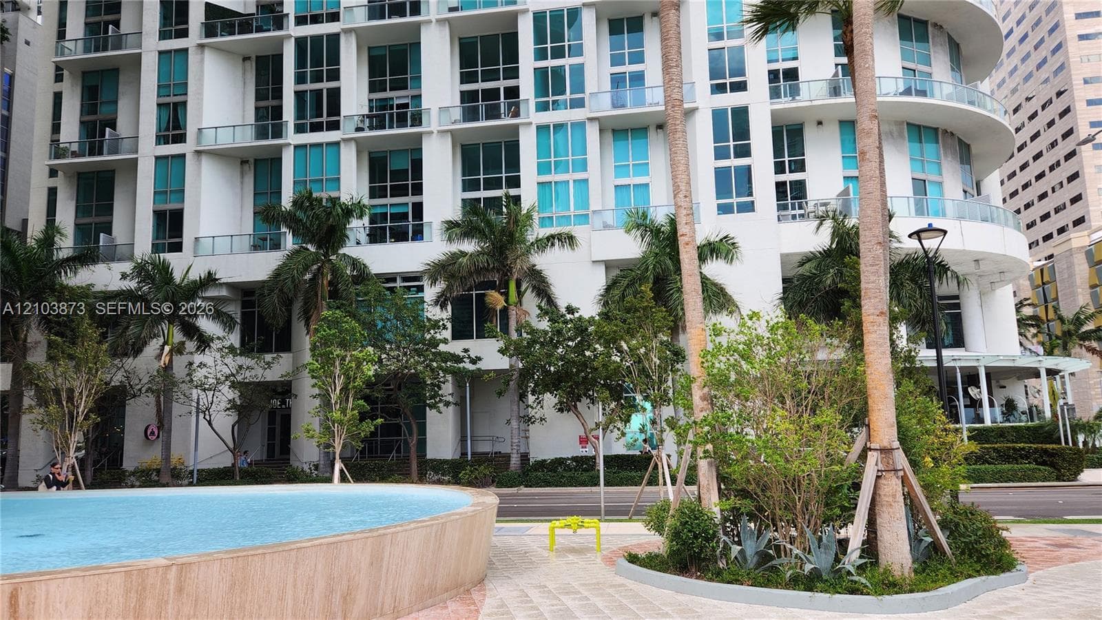 High-rise exterior featuring curved architectural design, floor-to-ceiling glass windows, private balconies, a circular water feature, and landscaped grounds with mature palm trees