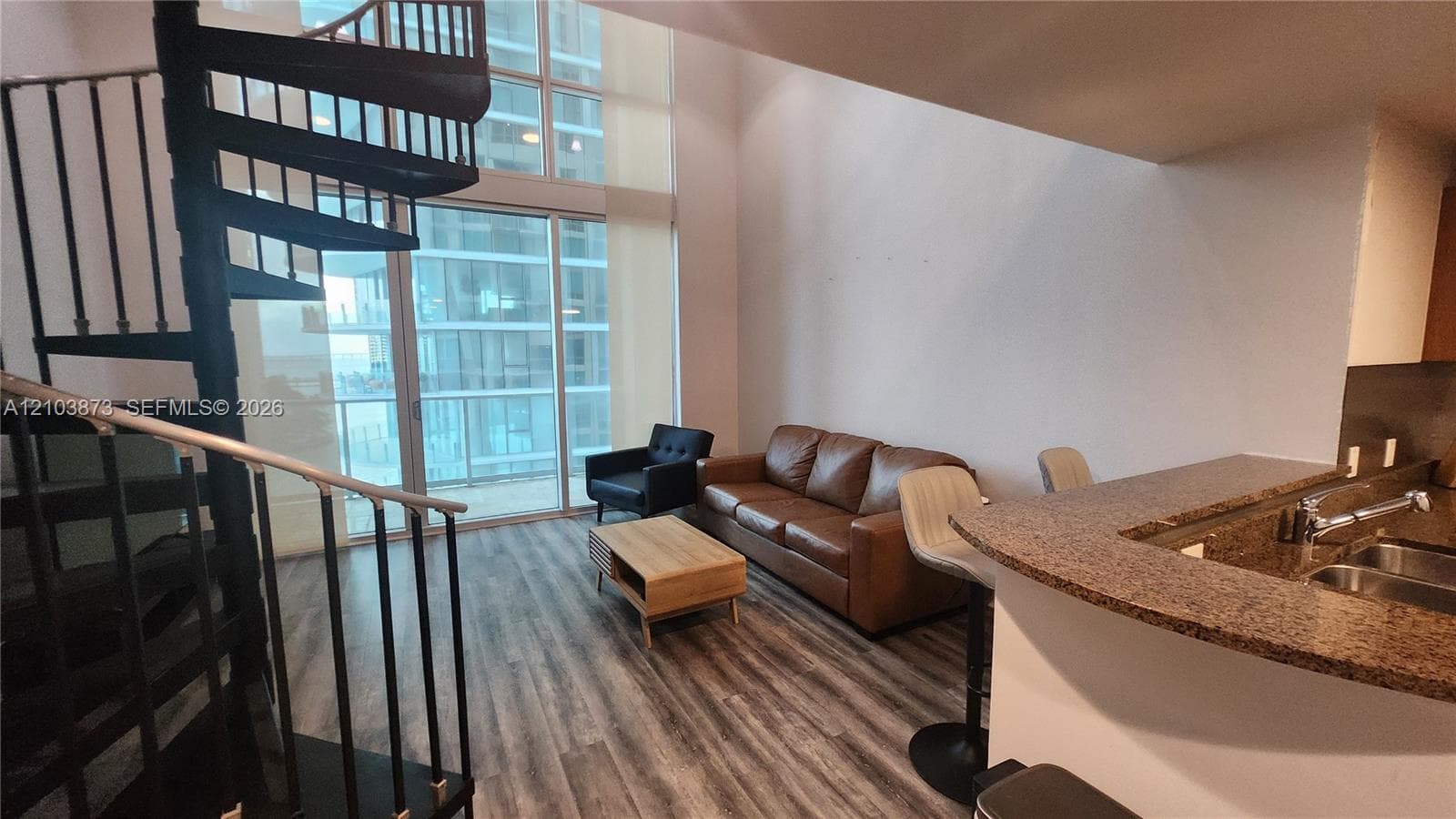 Double-height living area featuring floor-to-ceiling windows, a black metal spiral staircase, wood-finish flooring, and a kitchen peninsula with stone-finish countertops