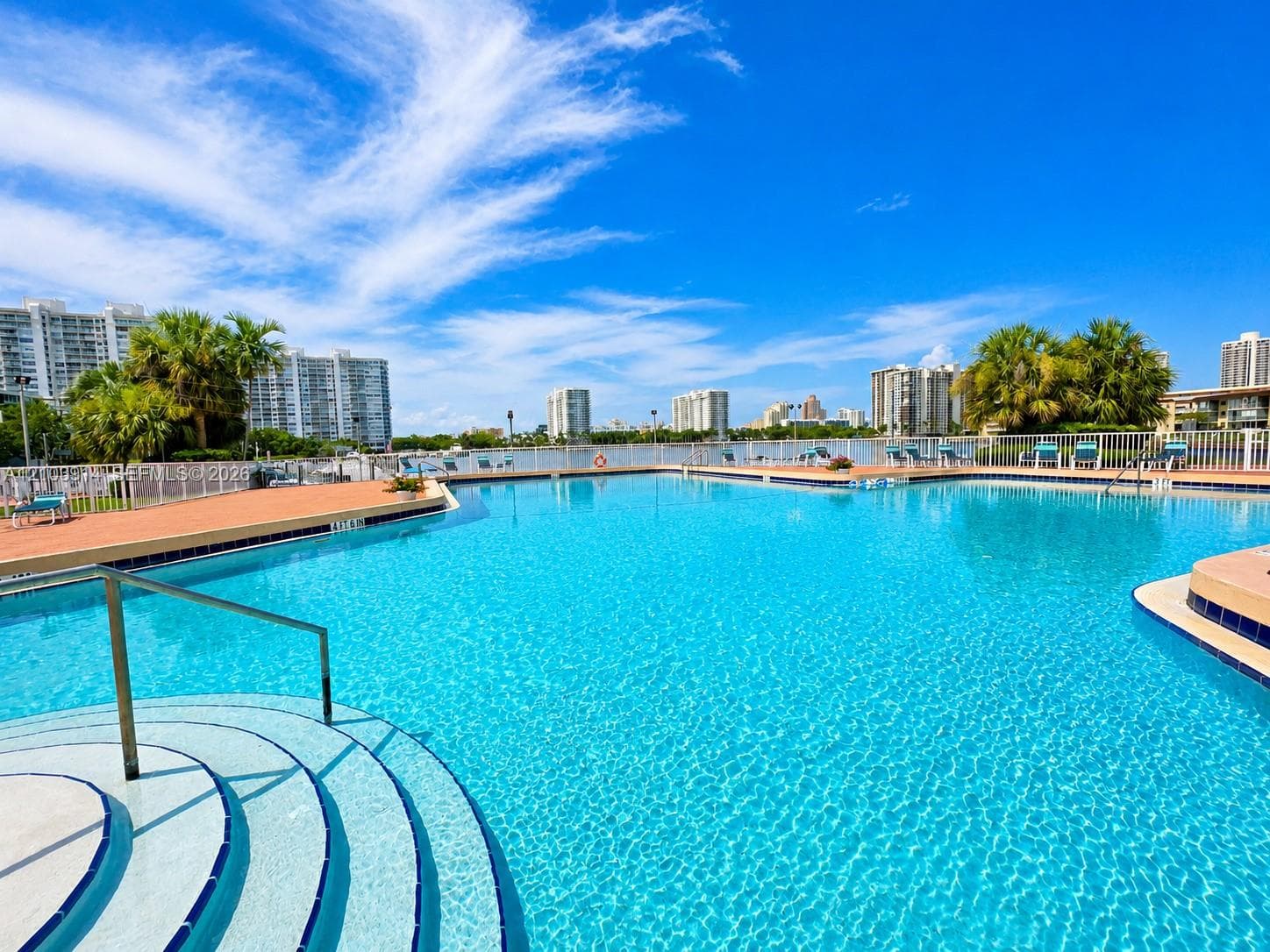 Swimming pool featuring submerged entry steps, a metal handrail, paved pool deck, and perimeter fencing
