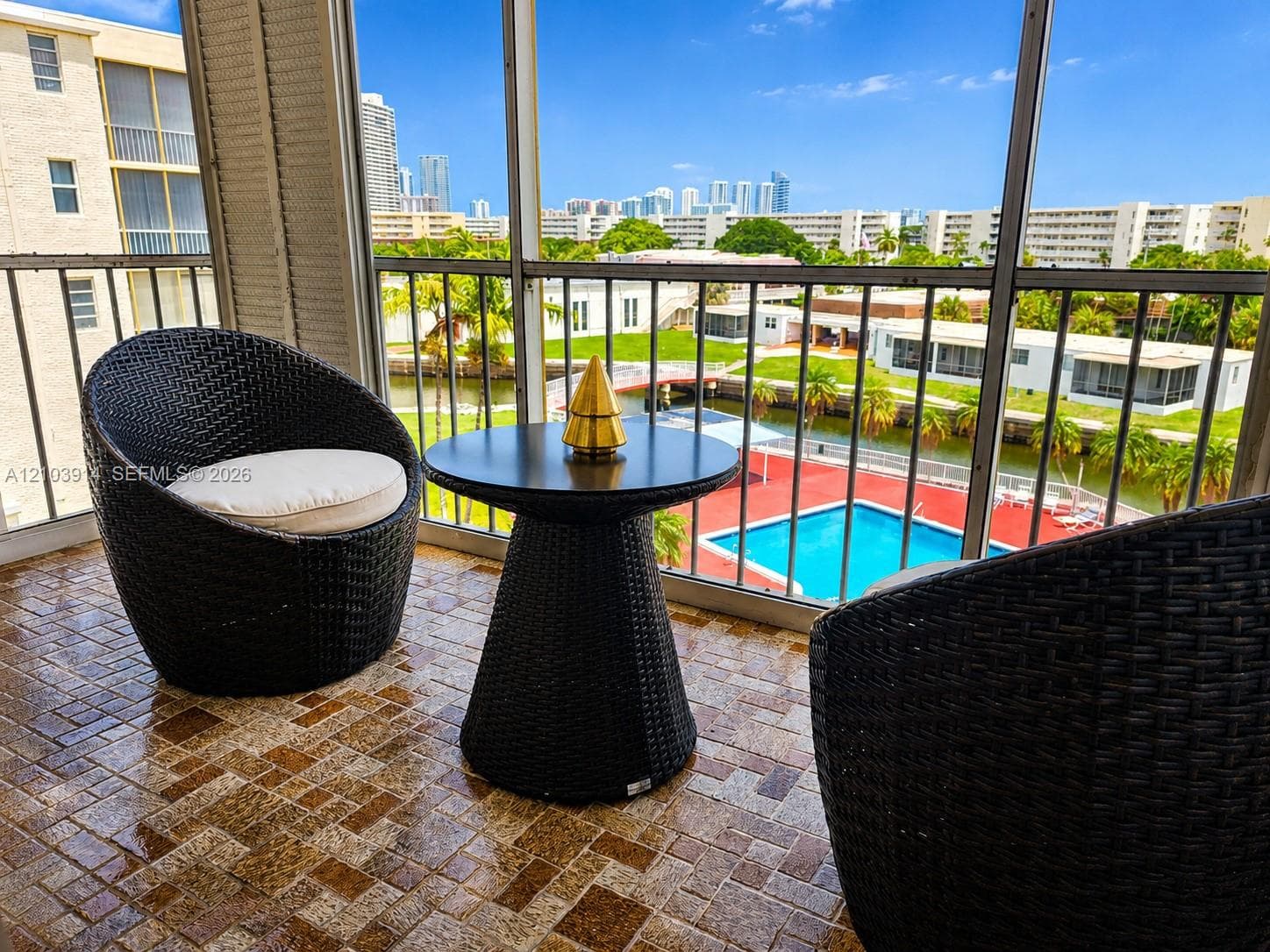 Screened balcony featuring tiled flooring and metal railing, overlooking an outdoor swimming pool, waterway, and city skyline