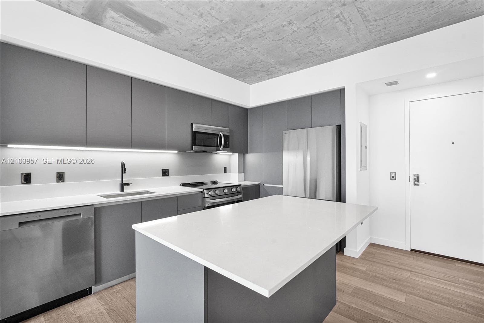 Modern kitchen featuring flat-panel cabinetry, stainless steel appliances, a center island with a light-toned countertop, a stainless steel sink with a dark-toned faucet, and wood-finish flooring