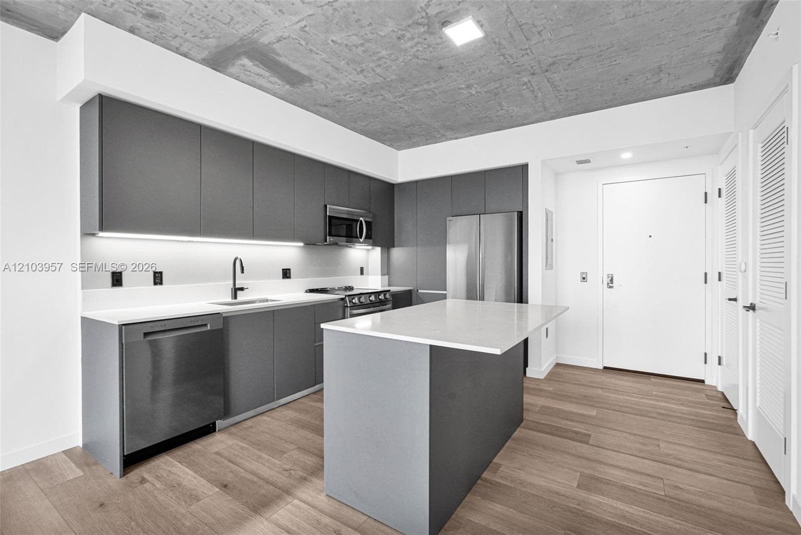 Kitchen featuring wood-finish flooring, an island with a light-toned countertop, stainless steel appliances, and sleek gray cabinetry with under-cabinet lighting