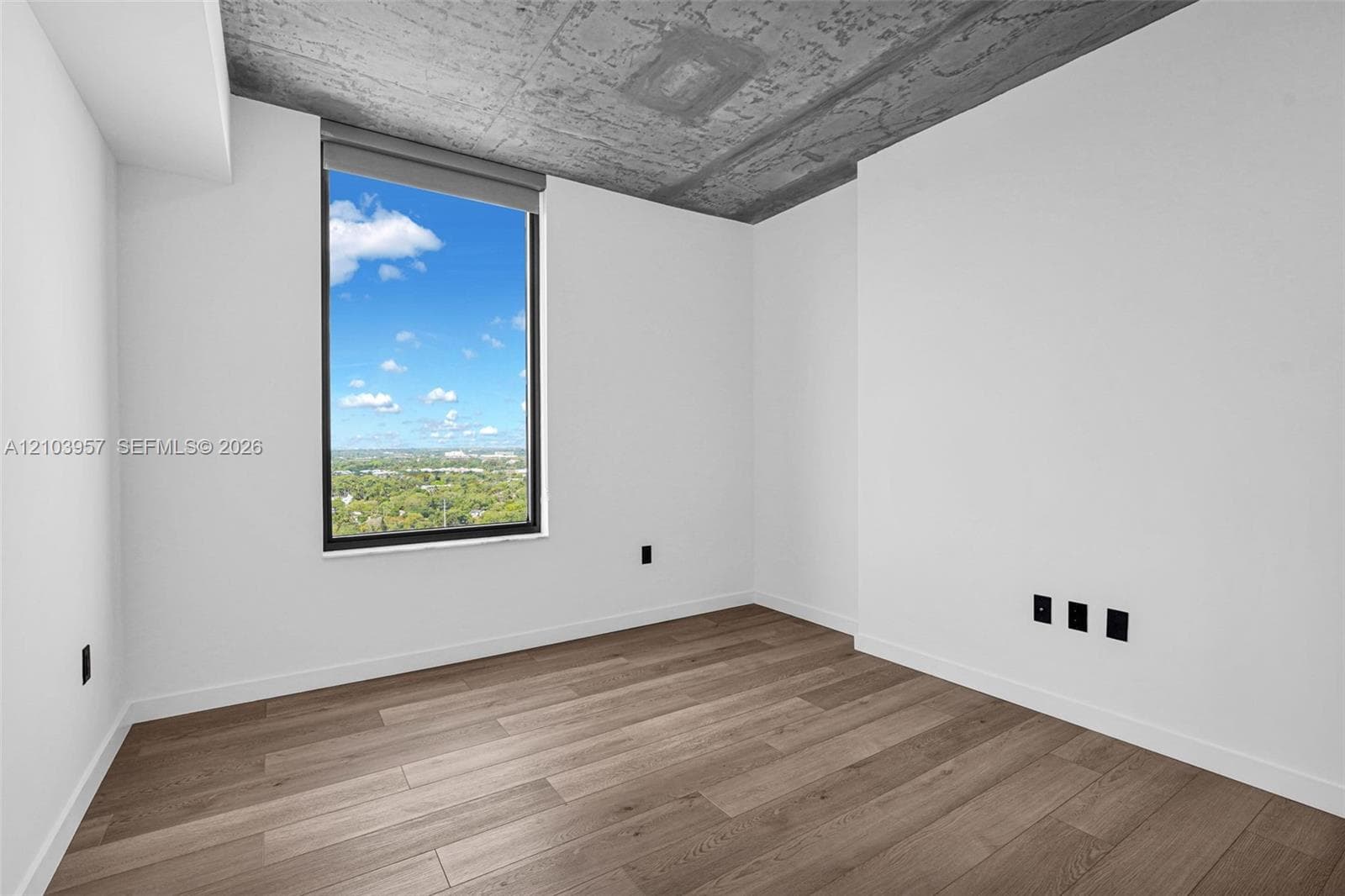Large window with a roller shade, wood-finish flooring, exposed concrete ceiling, and white walls