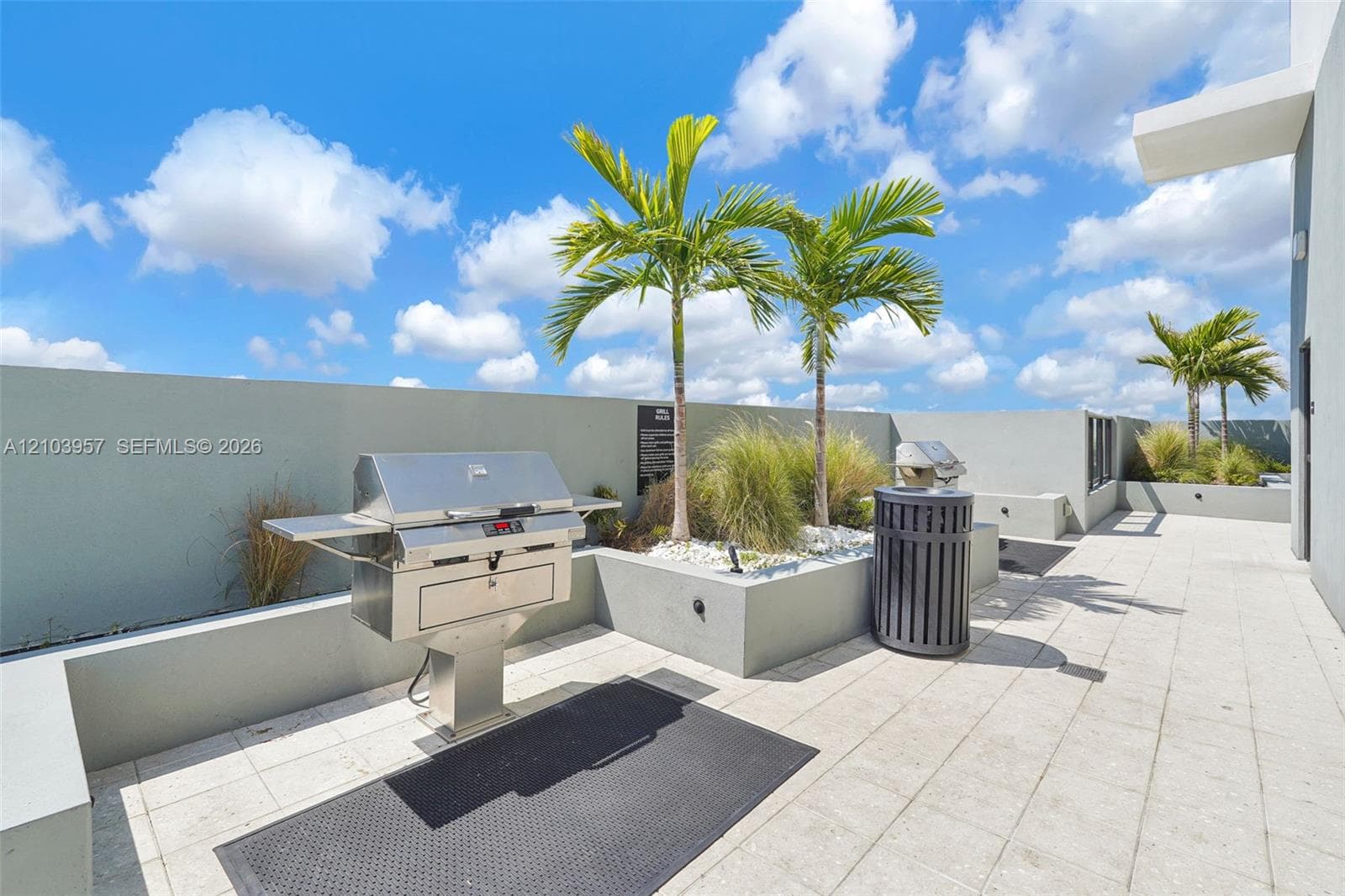 Rooftop patio featuring a built-in stainless steel grill, planter beds with palm trees, and light-toned tile flooring