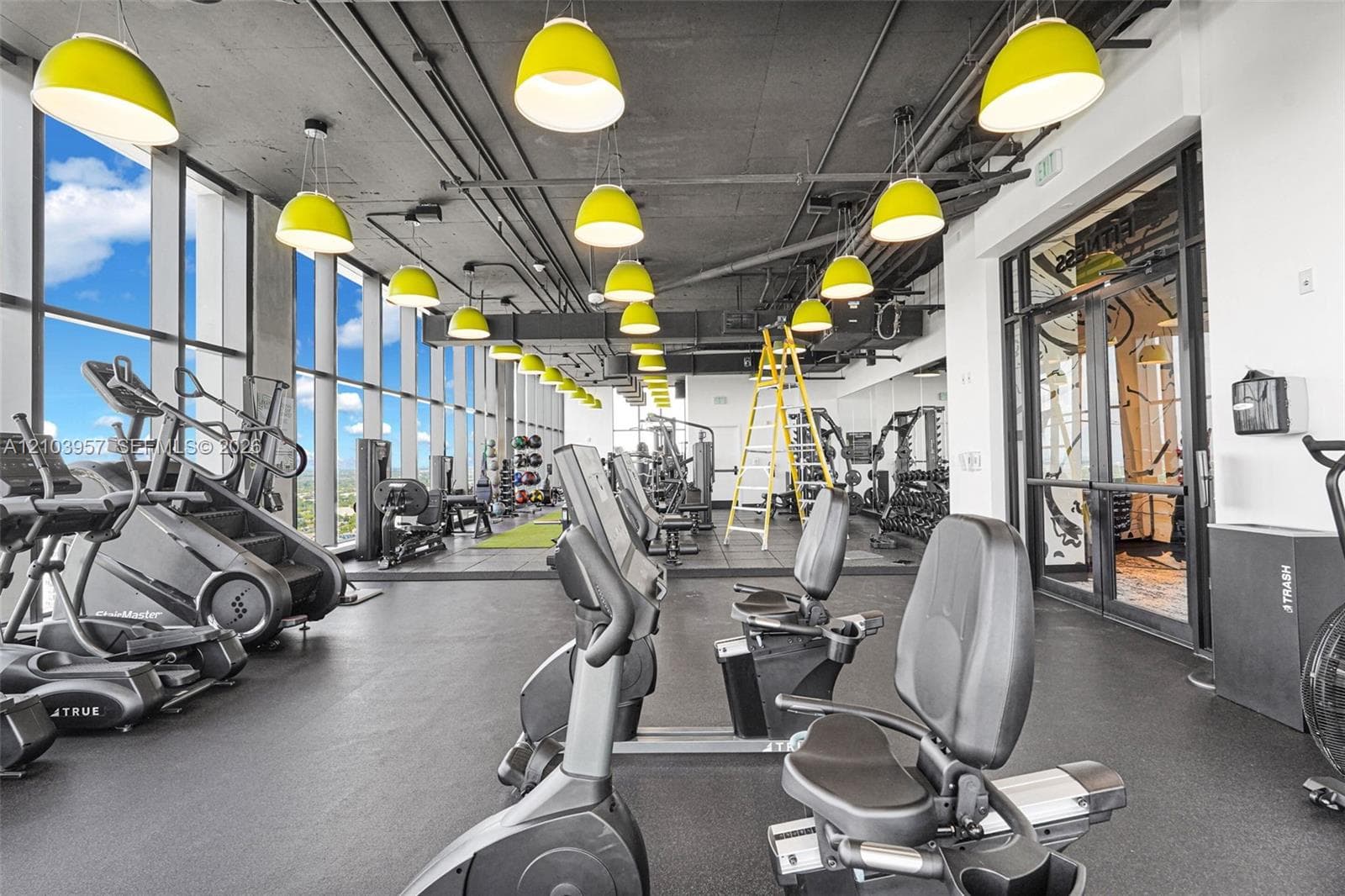 Floor-to-ceiling windows, exposed ceiling infrastructure, multiple pendant light fixtures, and dedicated exercise equipment
