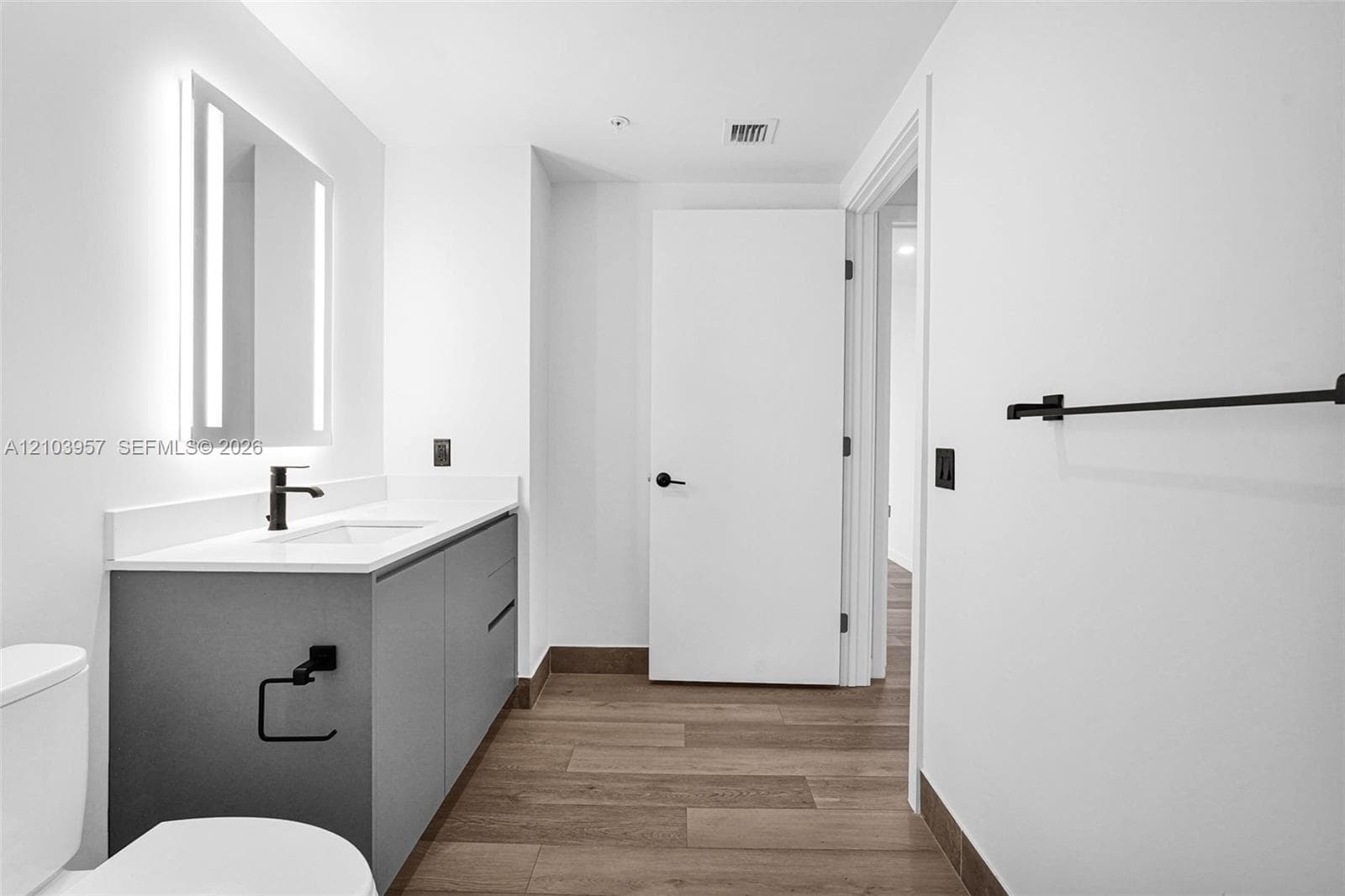 Contemporary bathroom featuring a floating vanity with a white countertop and integrated sink, an illuminated mirror, matte black fixtures, and wood-finish flooring