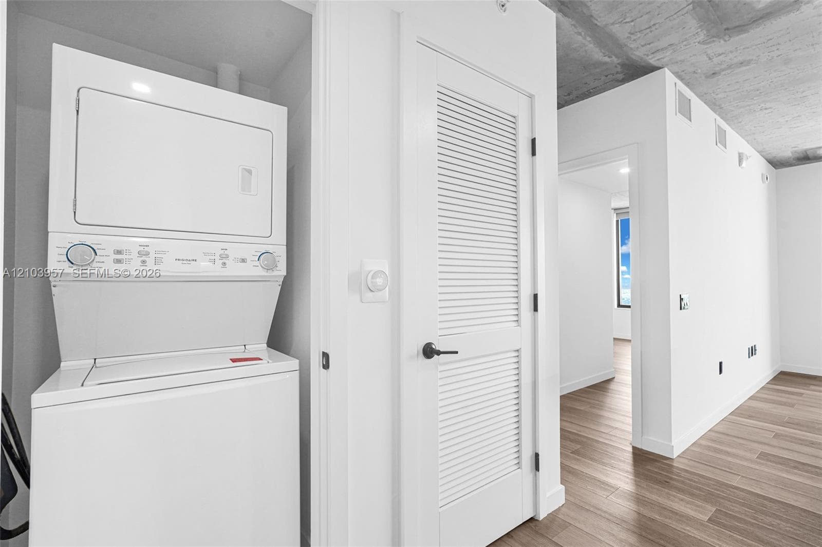 Stacked laundry appliances, louvered interior door, wood-finish flooring, exposed concrete ceiling, and white wall finishes