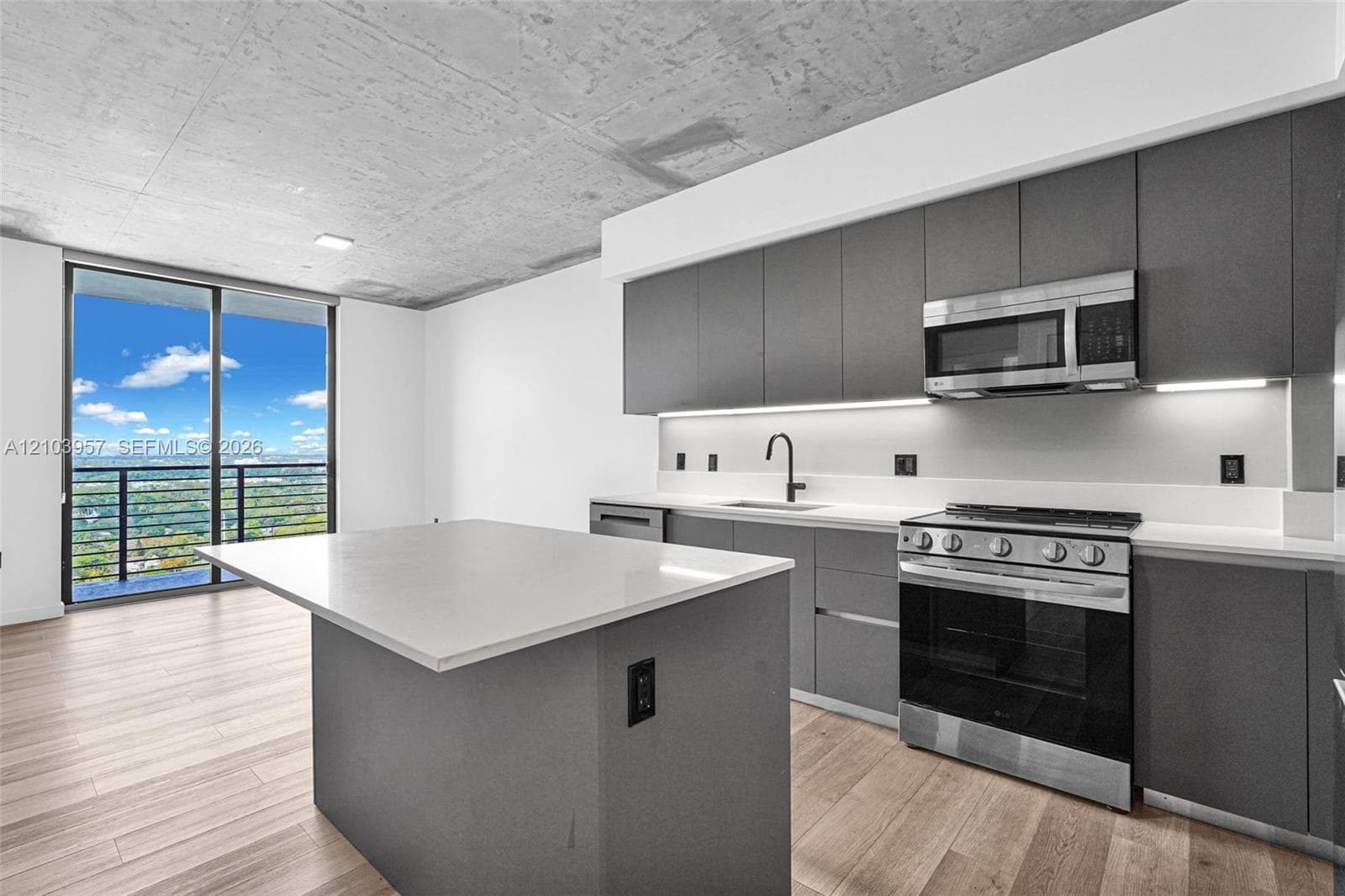Modern kitchen featuring an island with a light-toned countertop, stainless steel appliances, dark gray cabinetry, and wood-finish flooring