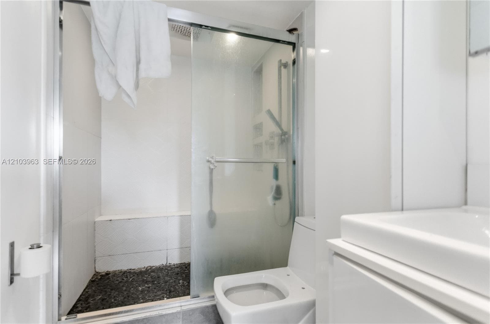 Shower enclosure with glass sliding doors, built-in tiled bench, stone-textured flooring, wall-mounted shower fixture, and white vanity with integrated basin