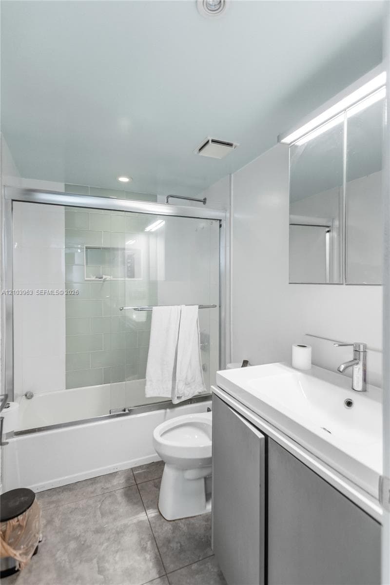 Bathroom featuring a vanity with an integrated sink, metal-finish faucet, medicine cabinet, stone-finish tile flooring, and a tub-shower combination with a sliding glass door and tiled surround