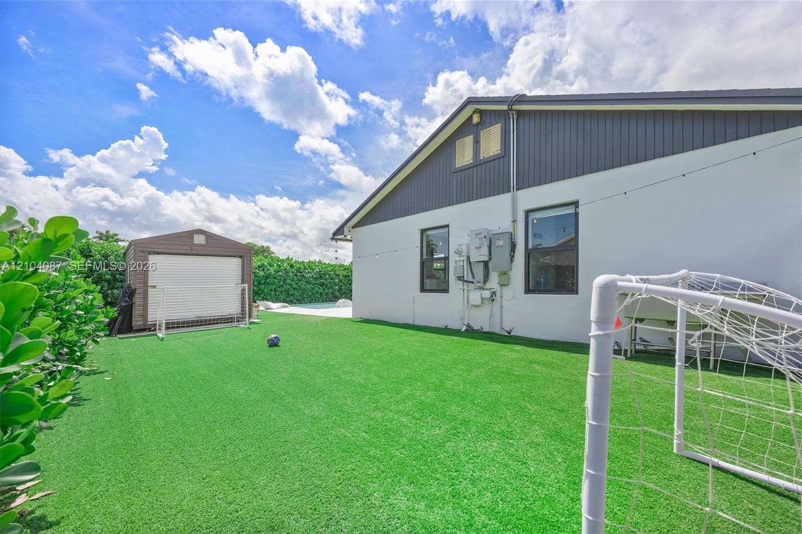 Expansive backyard featuring artificial turf, mature hedges, and a detached storage shed
