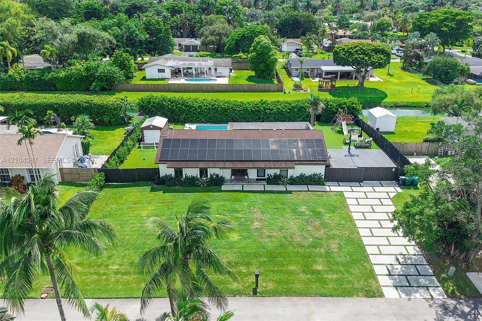 Paved driveway with rectangular stepping stones, mature palm trees, solar panel roof, detached outbuilding, and a fenced perimeter