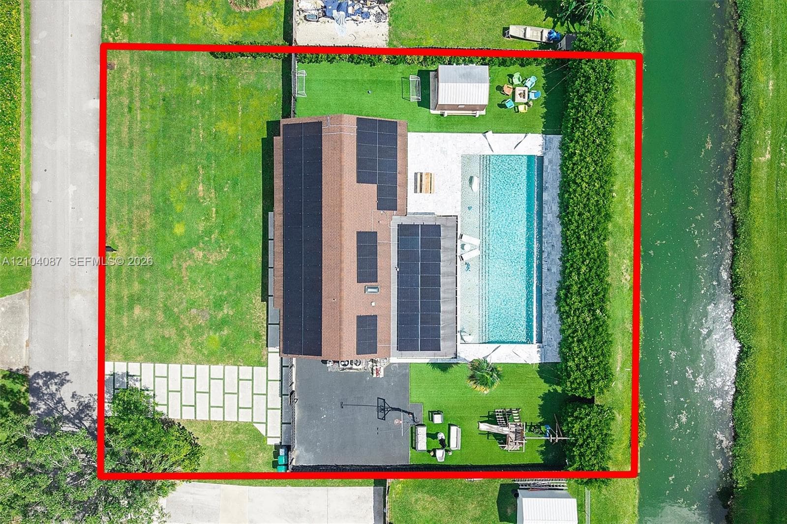 Rectangular swimming pool with light-colored decking, mature green landscaping, dark asphalt driveway, and a paved walkway