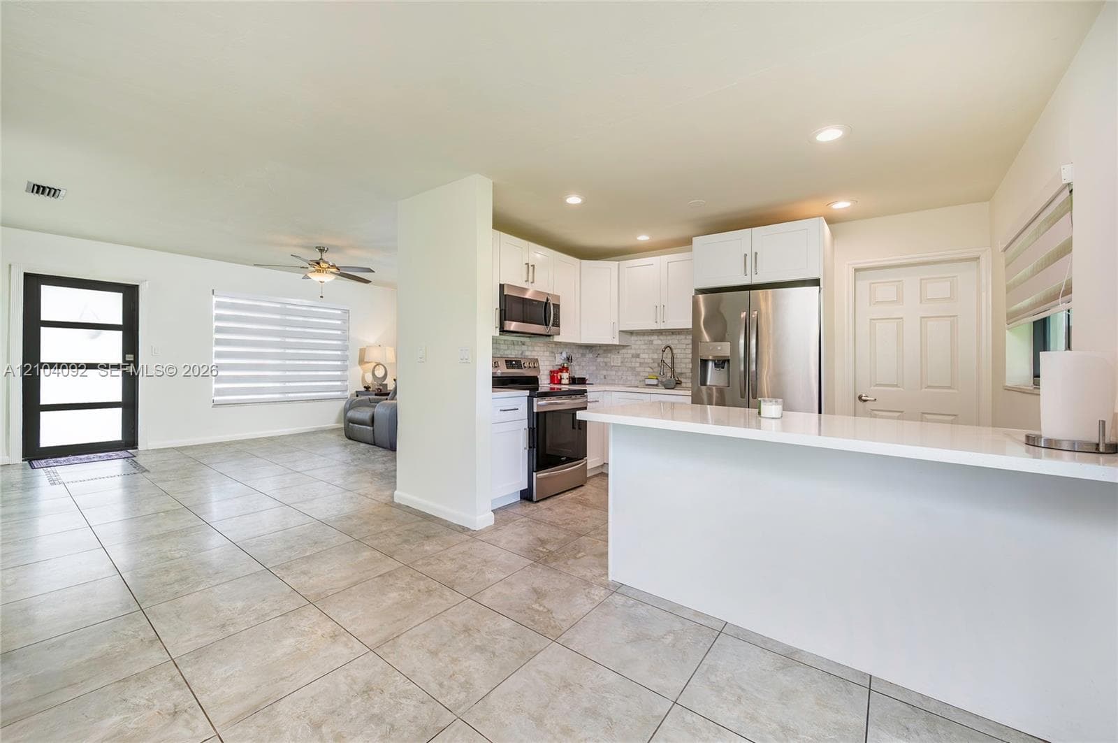 Kitchen featuring white shaker cabinetry, stainless steel appliances, stone-patterned backsplash, and a solid-surface breakfast bar, complemented by neutral tile flooring throughout