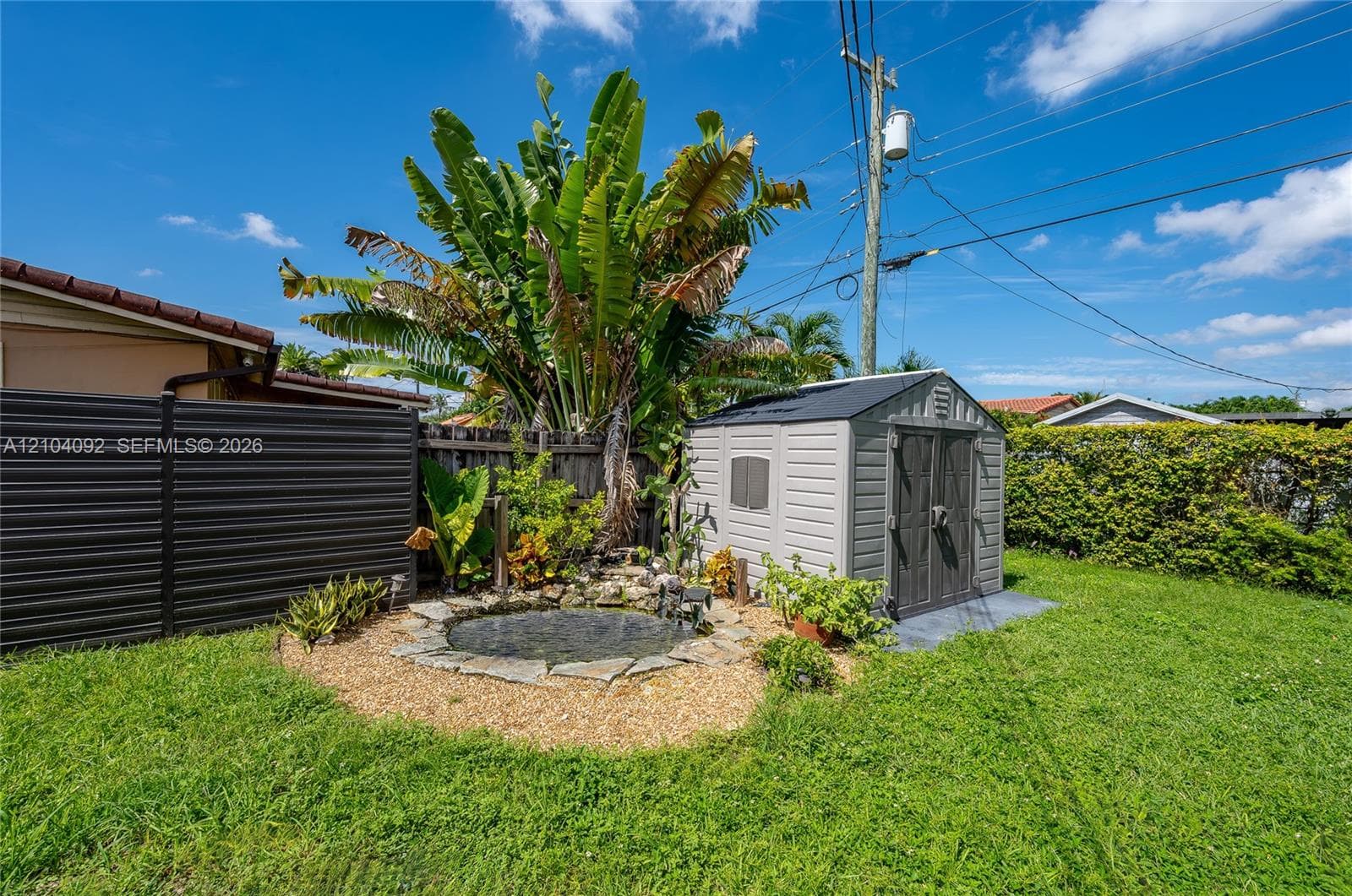 Backyard featuring a stone-edged pond, mature palm trees, a detached storage shed, metal privacy fencing, and green lawn space