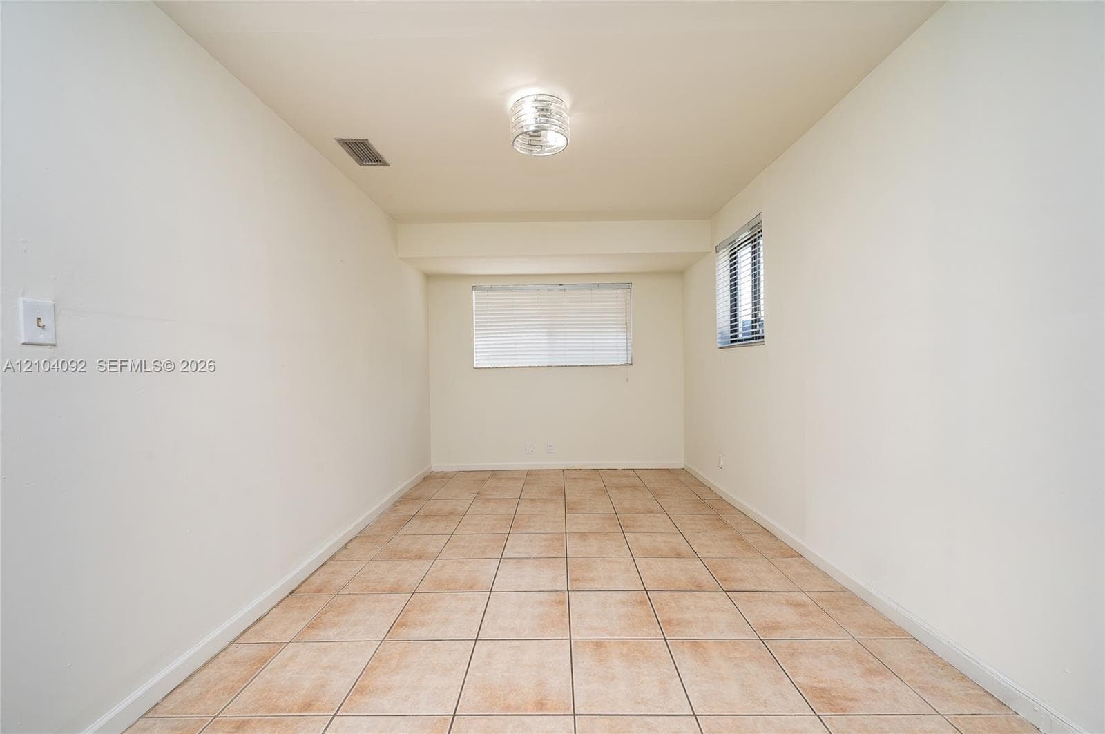 Neutral-toned interior space featuring light-colored ceramic tile flooring, neutral wall paint, a flush-mount circular ceiling fixture, and dual windows with horizontal blinds