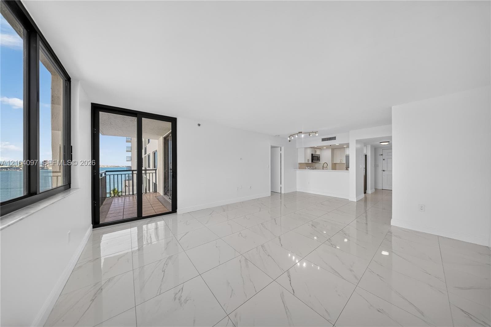 Open-concept living space featuring large-format polished tile flooring, floor-to-ceiling windows, sliding glass door access to a balcony, and white cabinetry in the kitchen area