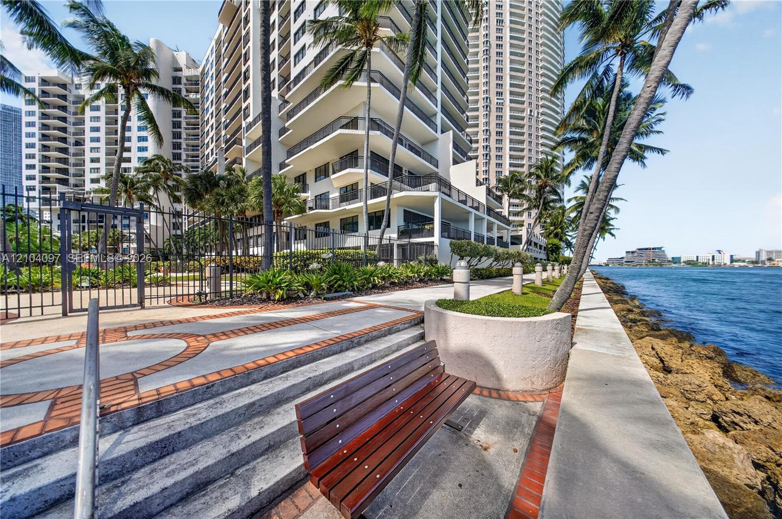 Waterfront promenade featuring a paved walkway with brick accents, concrete retaining walls, integrated landscaping, and metal perimeter fencing