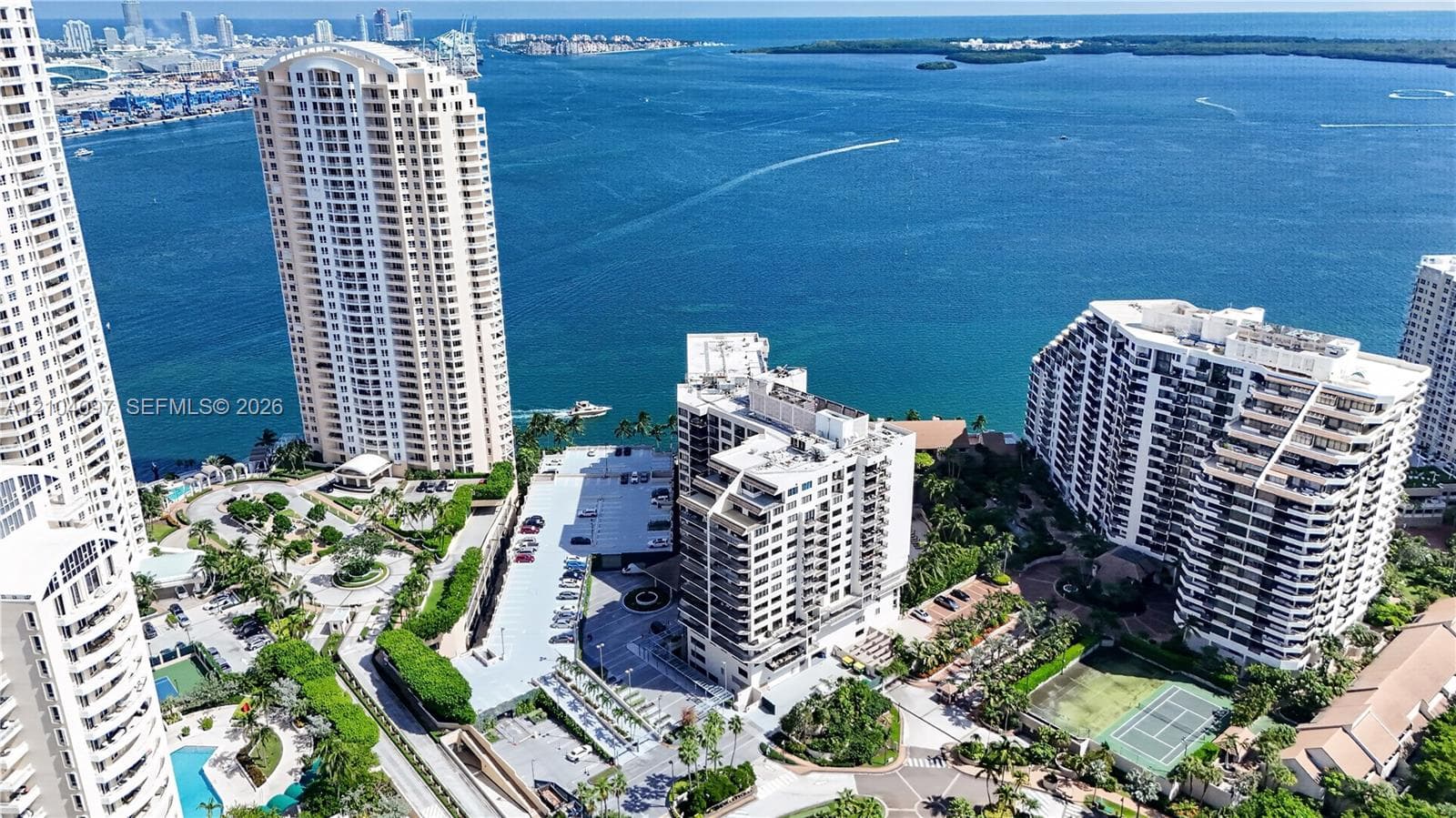 Aerial perspective featuring multiple high-rise residential buildings, waterfront access, hardscaped parking areas, and on-site tennis courts