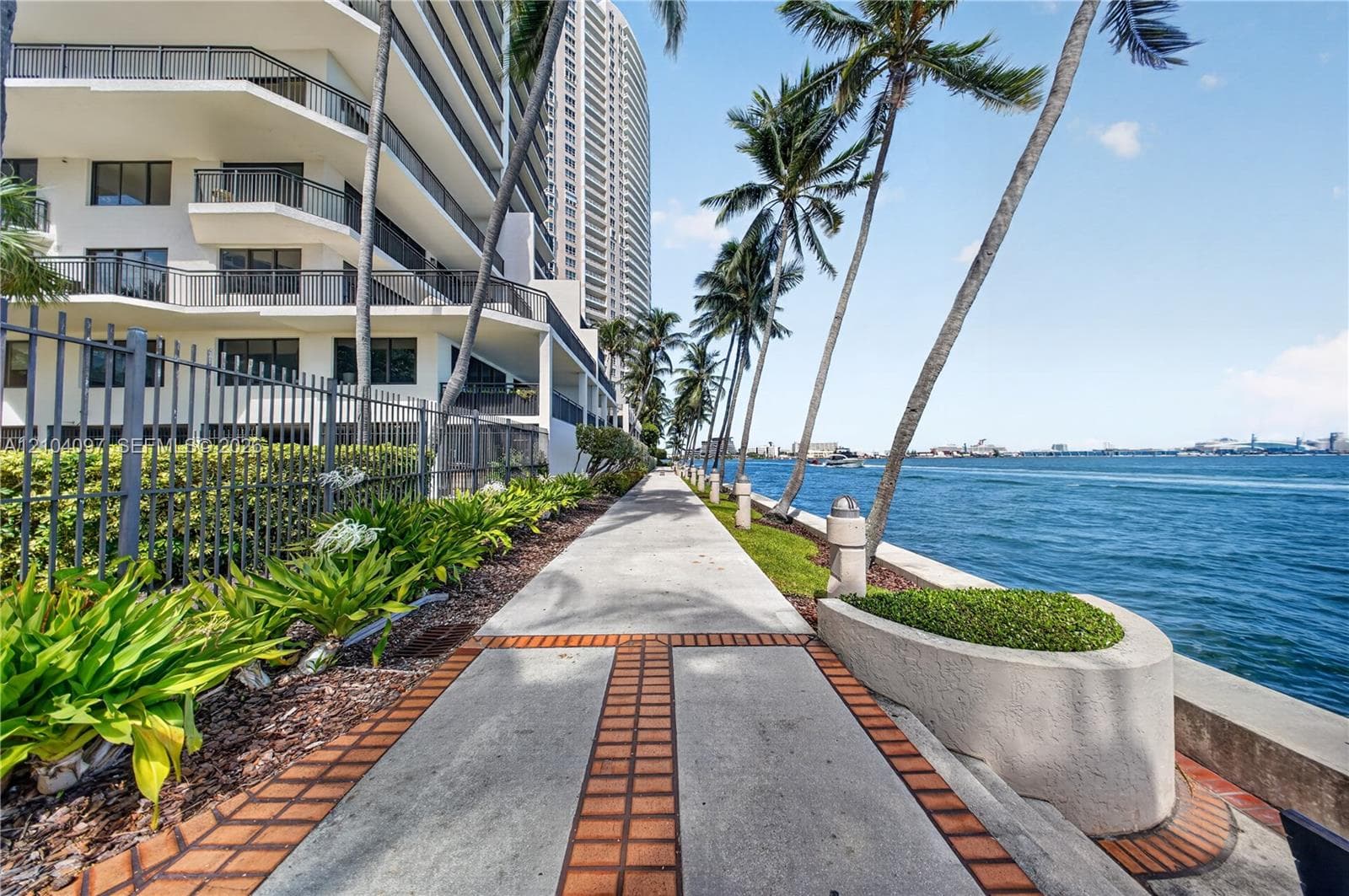 Concrete paved promenade featuring brick-accented inlay, waterfront bulkhead, native landscaping, and mature palm trees