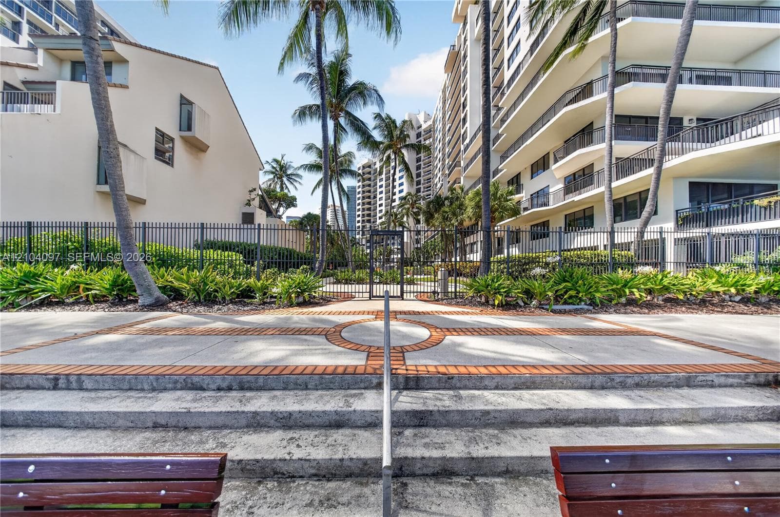 Concrete patio with inlaid brick detailing, metal perimeter fencing, tiered concrete steps with a central handrail, and mature palm trees