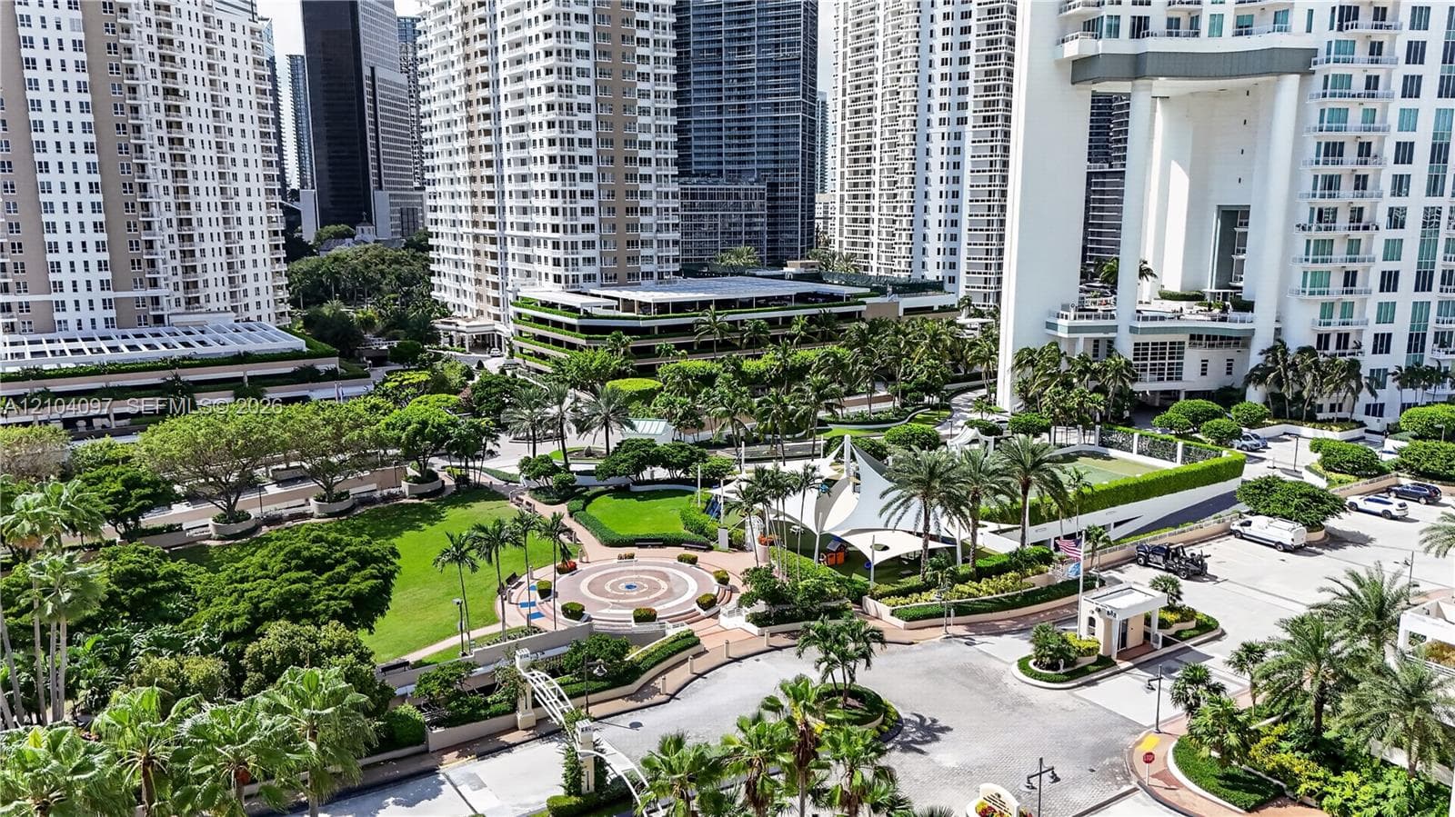 Elevated aerial perspective featuring a landscaped urban plaza, manicured green space, paved pathways, mature palm trees, and a tensile shade structure