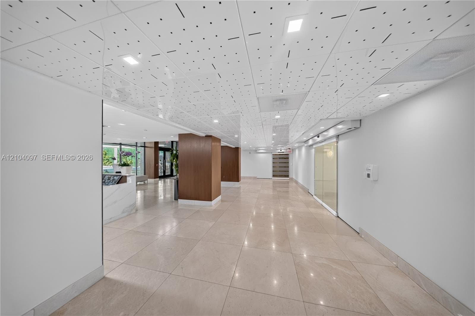 Expansive foyer featuring large-format tile flooring, a geometric patterned drop ceiling with recessed lighting, wood-veneer columns, and a reception desk with stone-finish surfacing