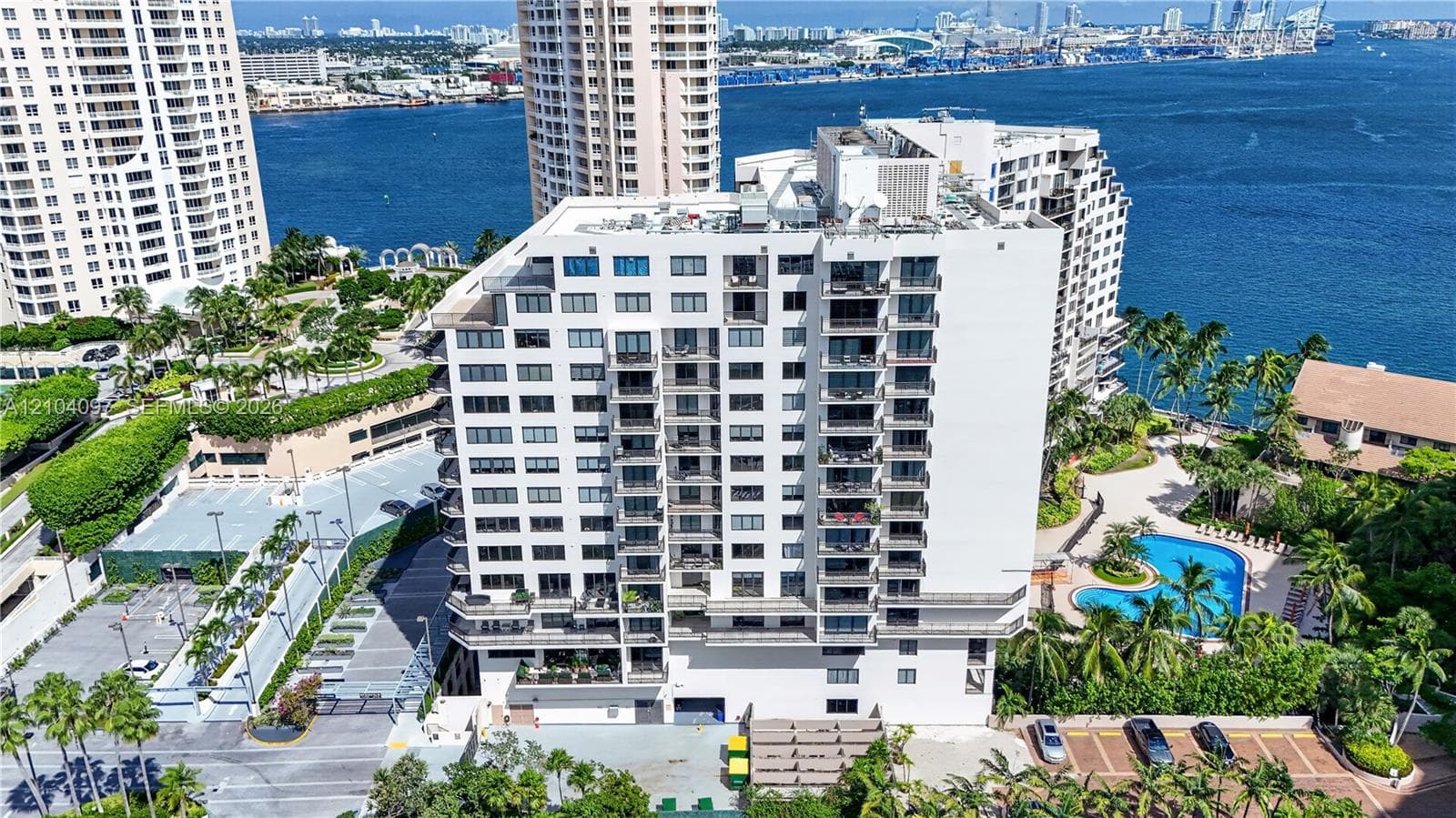 High-rise residential building featuring stacked private balconies, an on-site swimming pool, paved surface parking, and extensive tropical landscaping situated on the waterfront