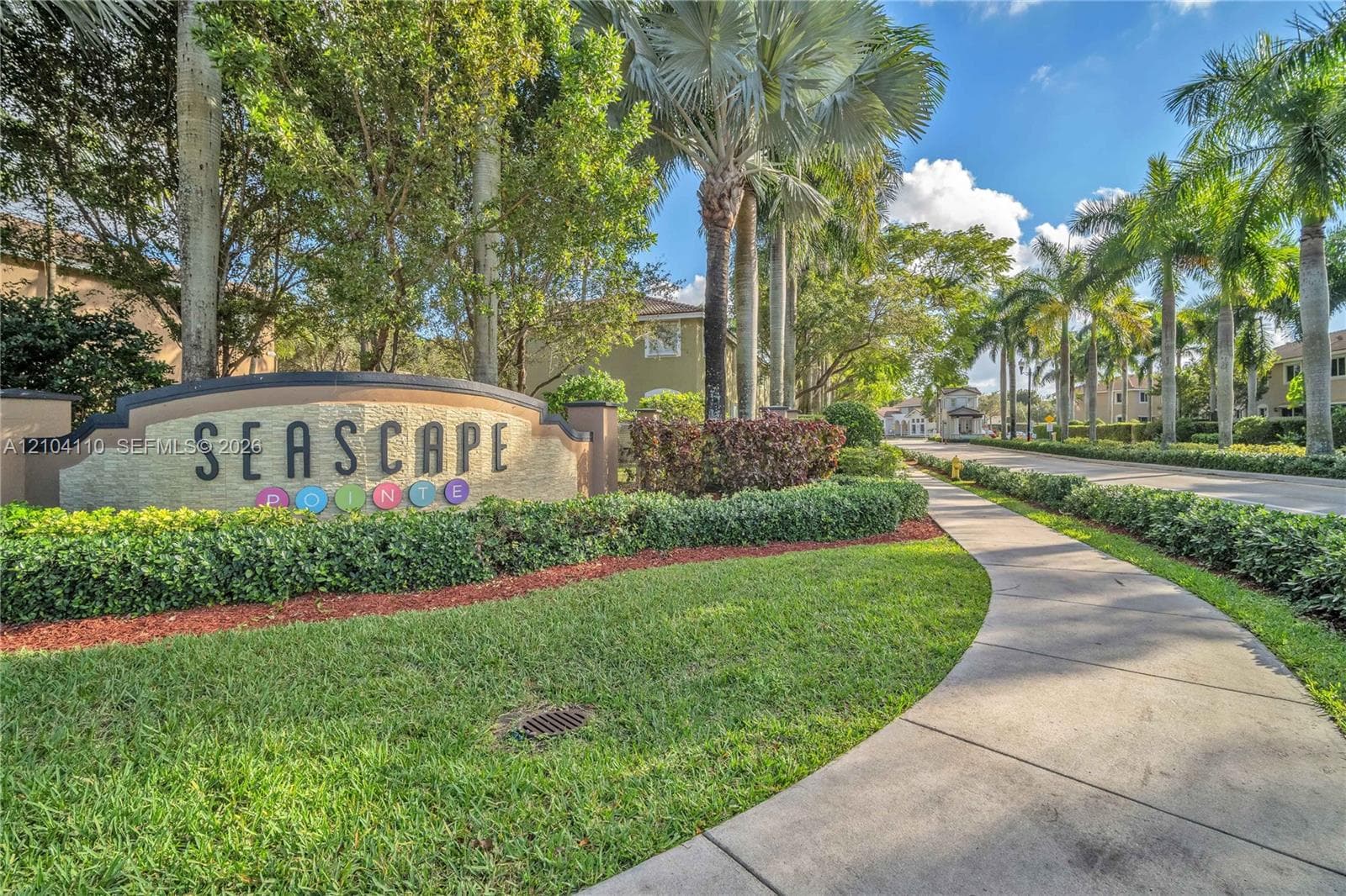 Entry monument with "SEASCAPE" lettering, curved concrete sidewalk, manicured lawn, established shrubbery, and mature palm trees lining the street