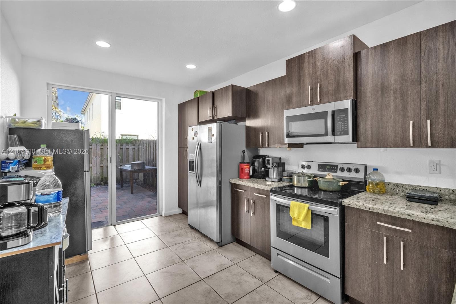 Kitchen featuring stainless steel appliances, wood-finish cabinetry, light-colored countertops, tile flooring, and a sliding glass door to a paved patio