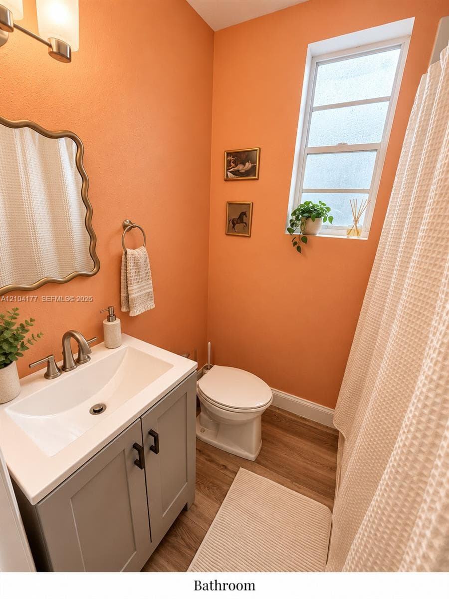 Vanity featuring a neutral-toned base, integrated sink, and polished metal hardware, paired with a decorative wall-mounted mirror, wood-finish flooring, and a window with frosted glass