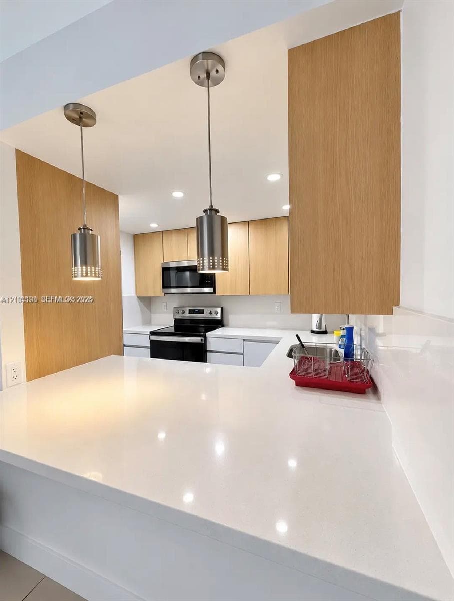 Kitchen featuring flat-panel wood-finish cabinetry, white solid-surface countertops, stainless steel appliances, and dual pendant lighting
