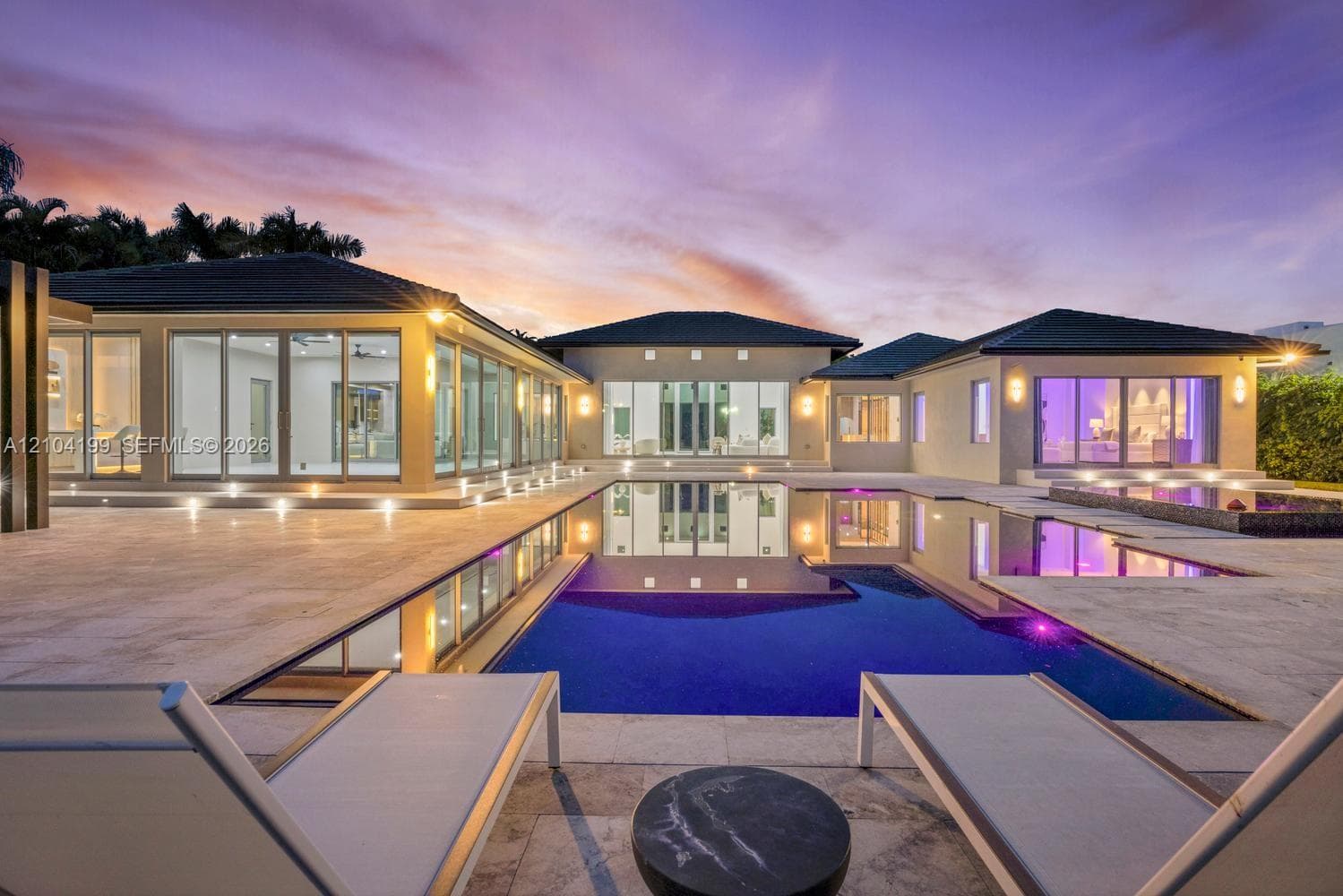 Single-story residence featuring an expansive stone-paved patio, an infinity-edge swimming pool, extensive floor-to-ceiling glass wall panels, and recessed exterior lighting