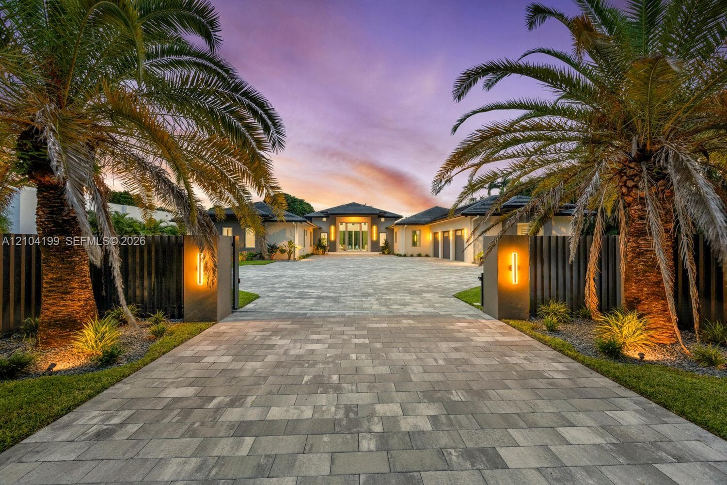 Paver stone driveway featuring an gated entrance with vertical fencing, mature palm trees, and wall-mounted exterior lighting
