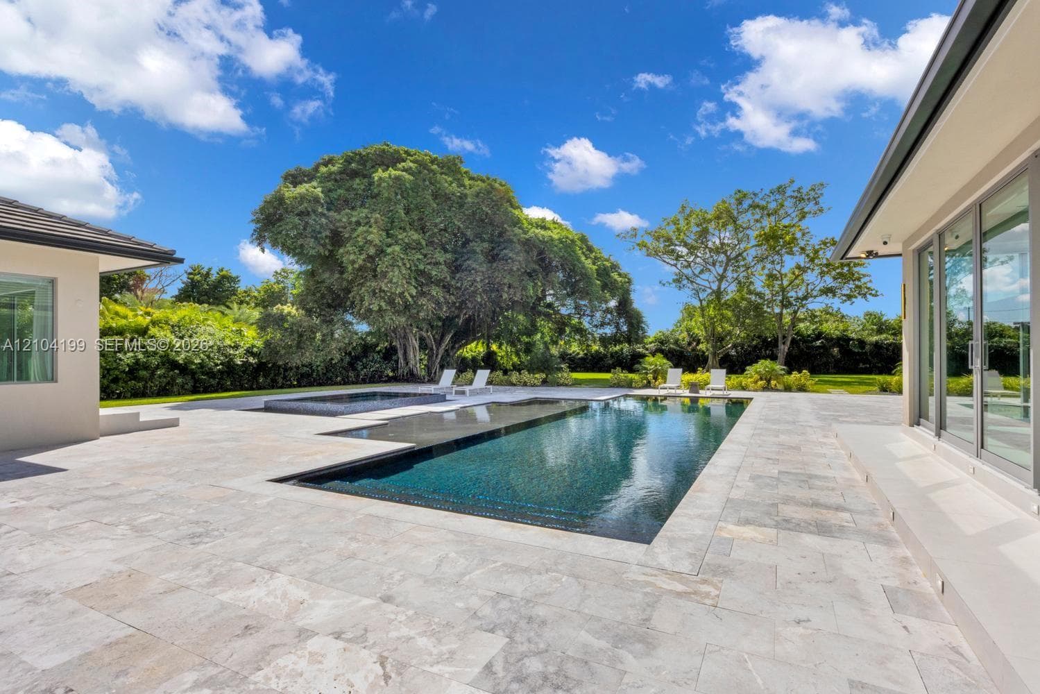 Expansive stone patio featuring a multi-level swimming pool with a raised spa, built-in covered pergola, and floor-to-ceiling glass wall panels