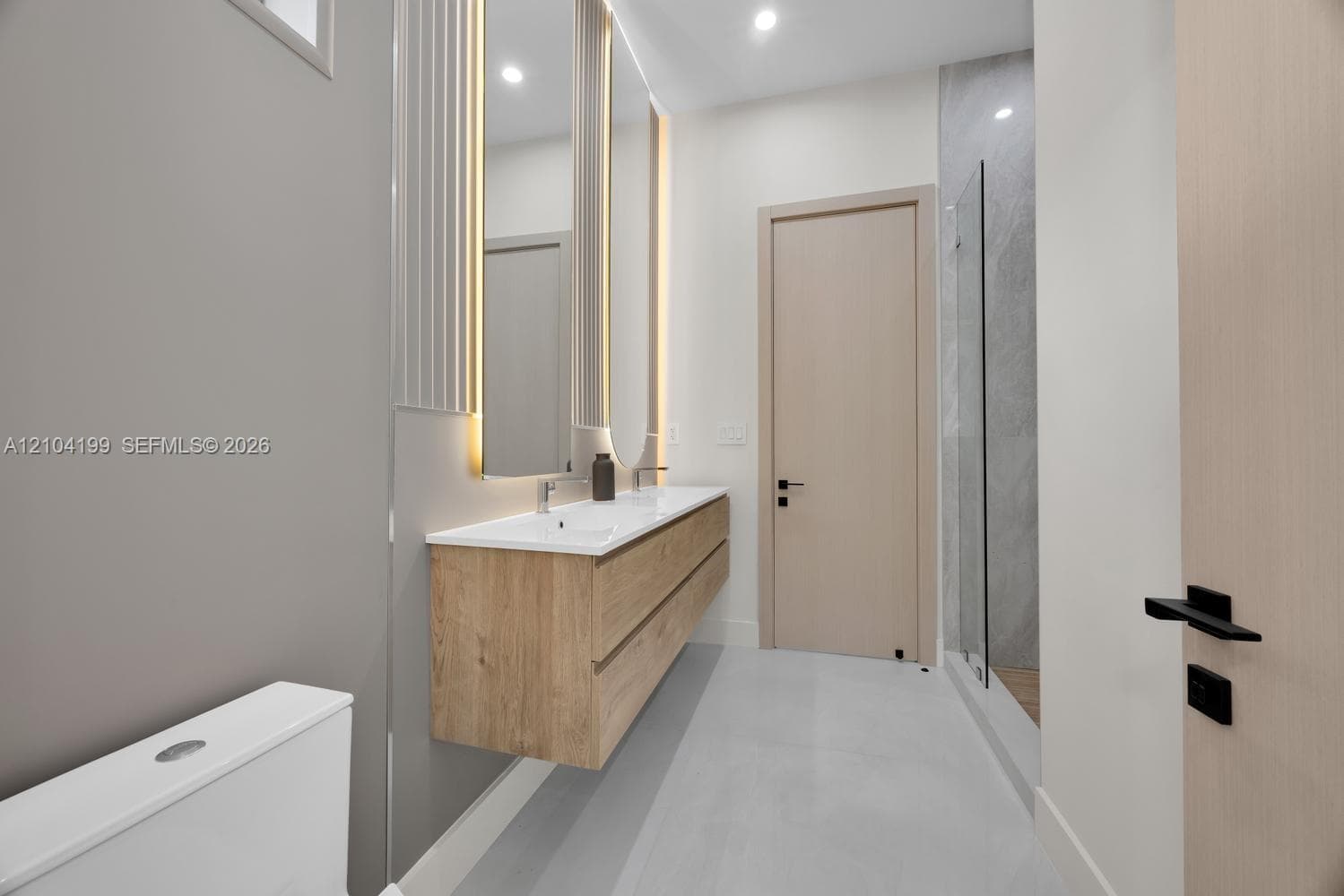 Bathroom featuring a floating vanity with a white countertop, an arched LED-backlit mirror, large-format grey tile walling, and a walk-in shower with a glass enclosure