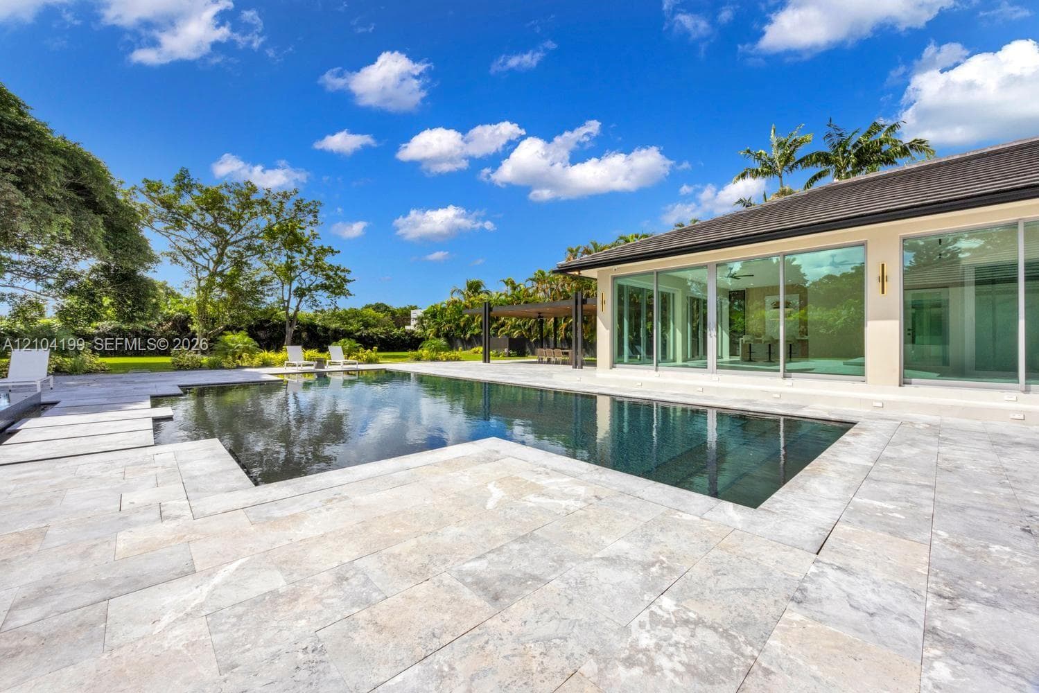Spacious outdoor living area featuring extensive stone paver hardscaping, an in-ground swimming pool, a covered pavilion, and floor-to-ceiling glass panel doors