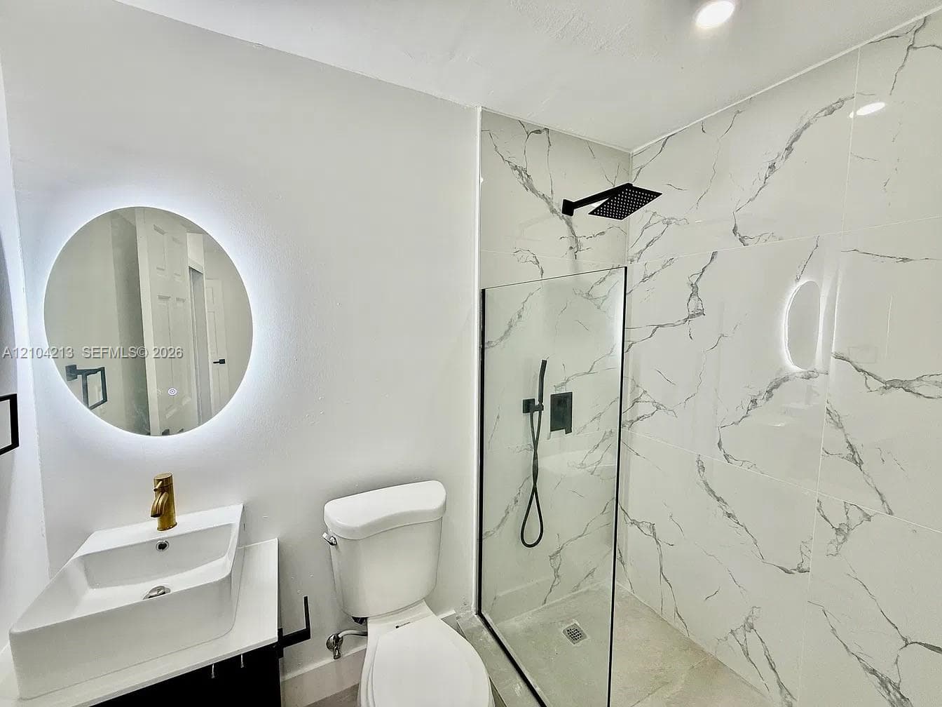 Modern bathroom featuring a rectangular vessel sink with gold-finish faucet, an illuminated circular mirror, and a walk-in shower with black-finish fixtures and patterned wall tile
