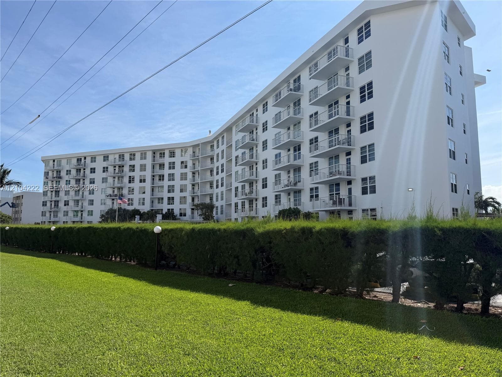 Multi-story residential building featuring white stucco facade, stacked private balconies, and landscaped hedge perimeter bordering a grass lawn
