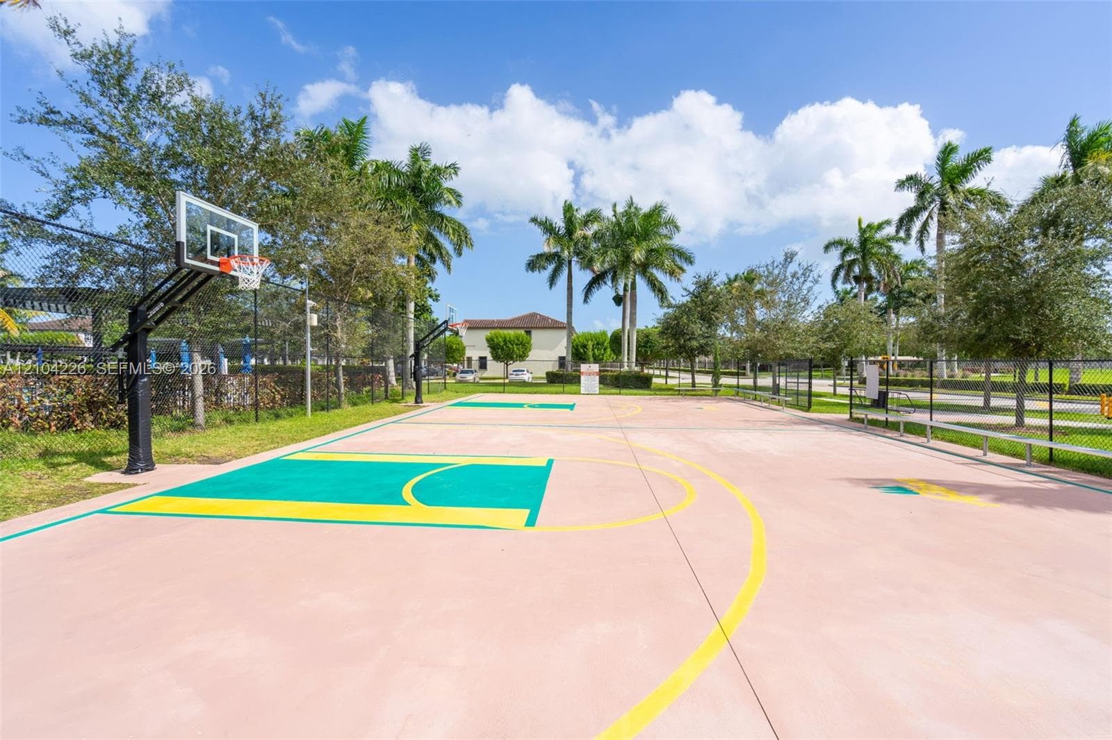 Outdoor basketball court with a multi-colored paved surface, steel hoops, and perimeter chain-link fencing, surrounded by mature palm trees and manicured landscaping