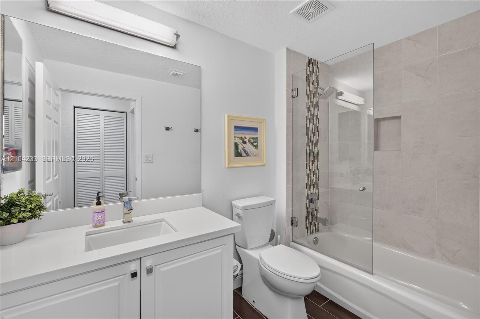 Bathroom featuring a vanity with a white countertop and rectangular undermount sink, a large wall-mounted mirror, a tub-shower combination with glass paneling, a vertical tile accent border, and recessed wall shelving