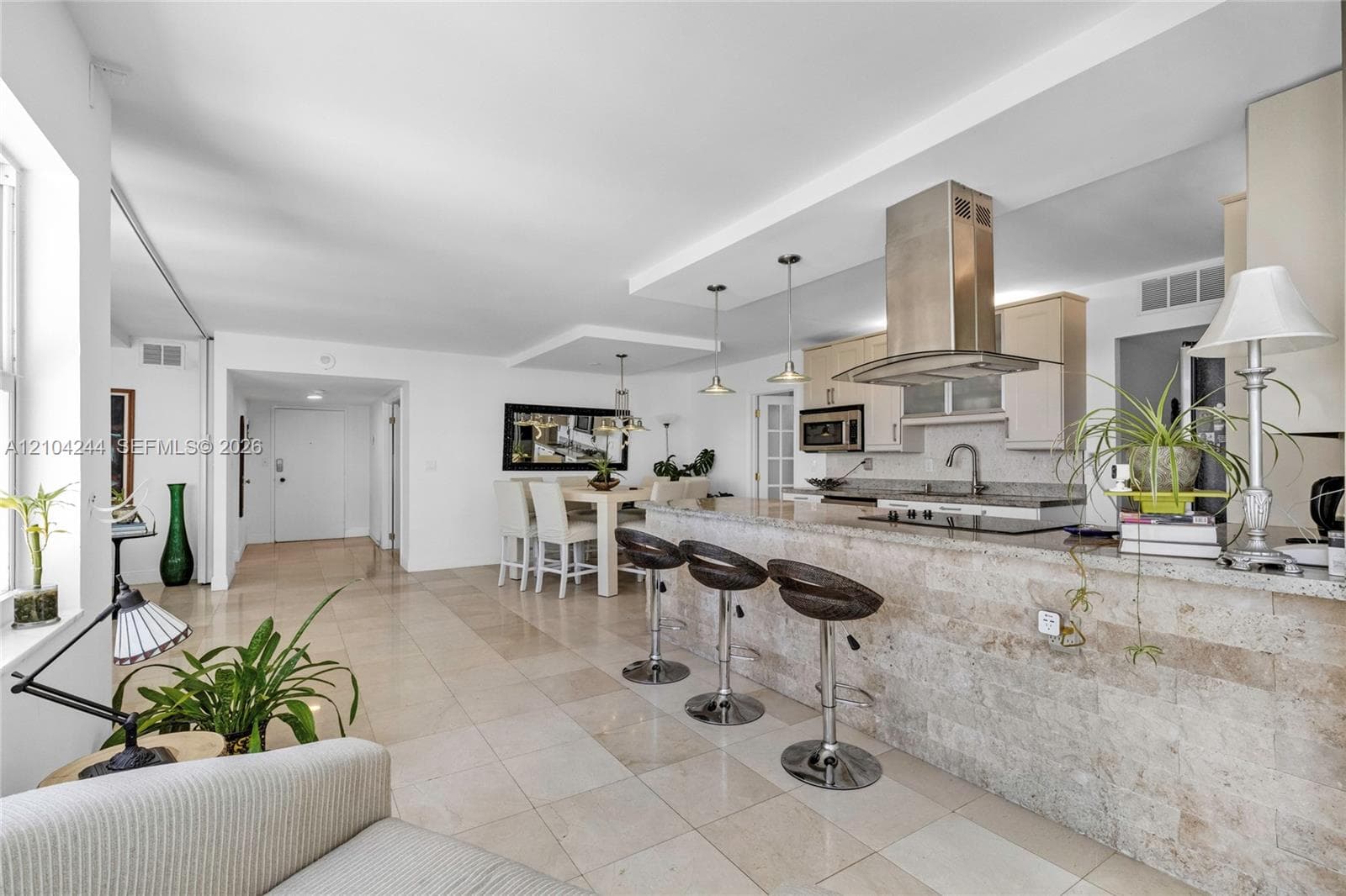 Open-concept kitchen featuring stone-clad breakfast bar, stainless steel chimney-style range hood, light-toned cabinetry, and tile flooring